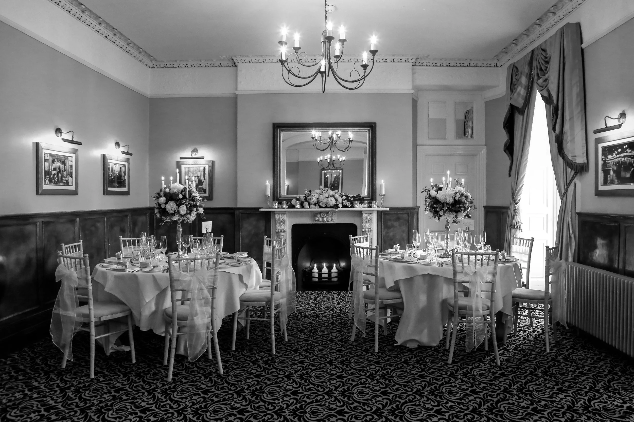 Elegant dining room decorated for a formal event with round tables, floral centerpieces, and tall candles, featuring a large mirror above a fireplace, ornate crown molding, and heavy draped curtains.