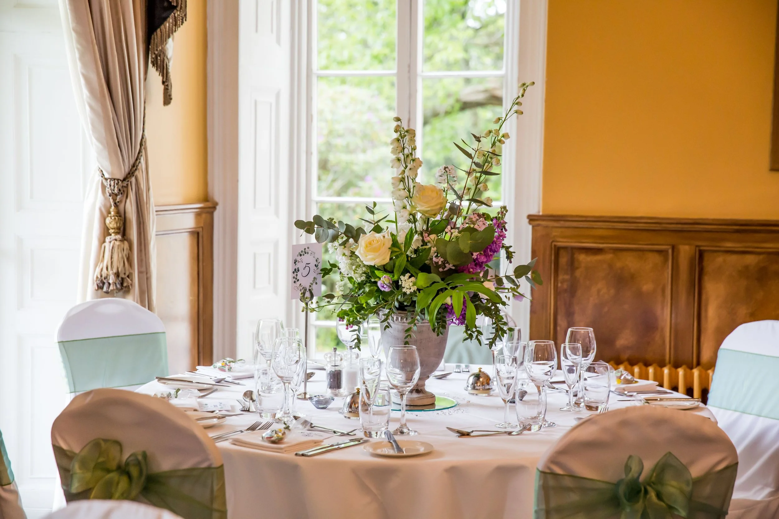 Elegant dining table set with champagne glasses, silverware, and a large floral centerpiece in a well-lit room with large windows and yellow walls.