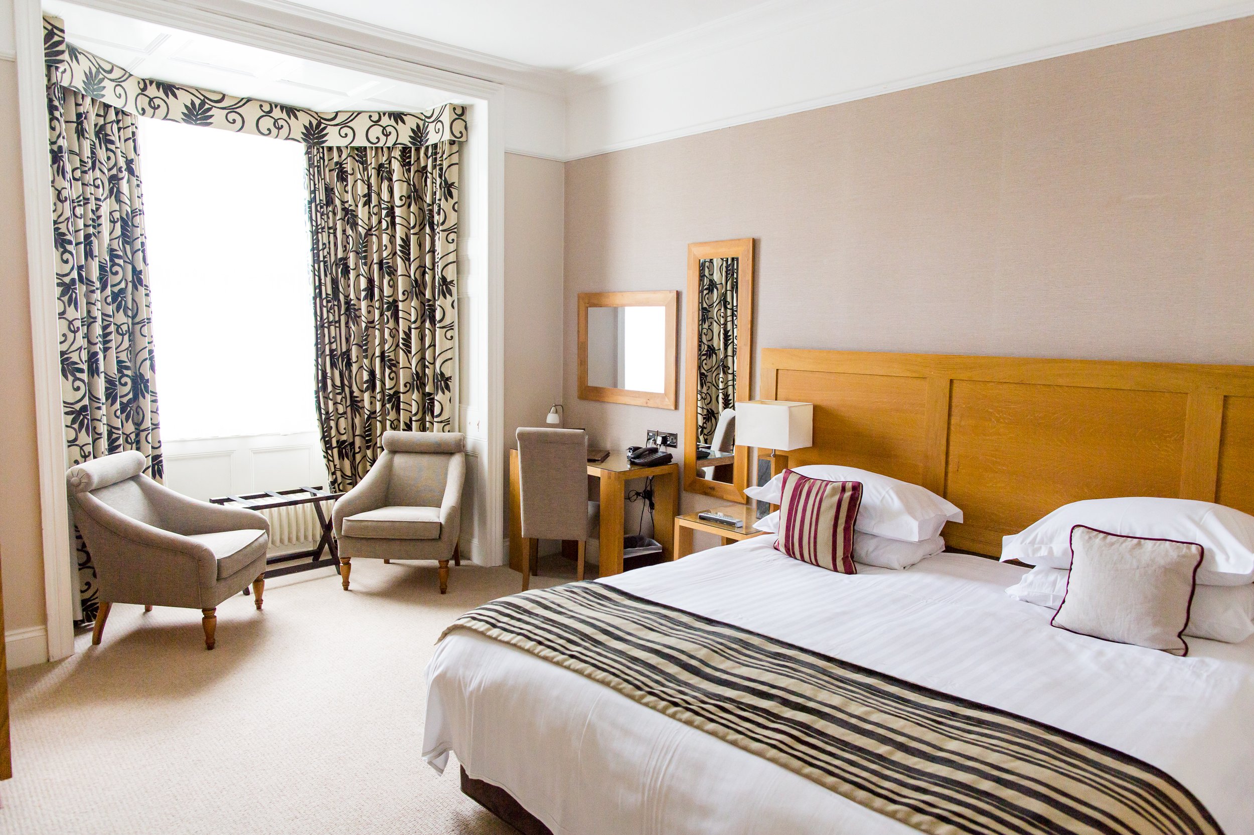 Hotel room with a large bed, wooden headboard, striped runner, pillows, bedside lamps, a marble-topped desk with a chair, full-length mirror, two armchairs near a window with patterned curtains, and a small table.