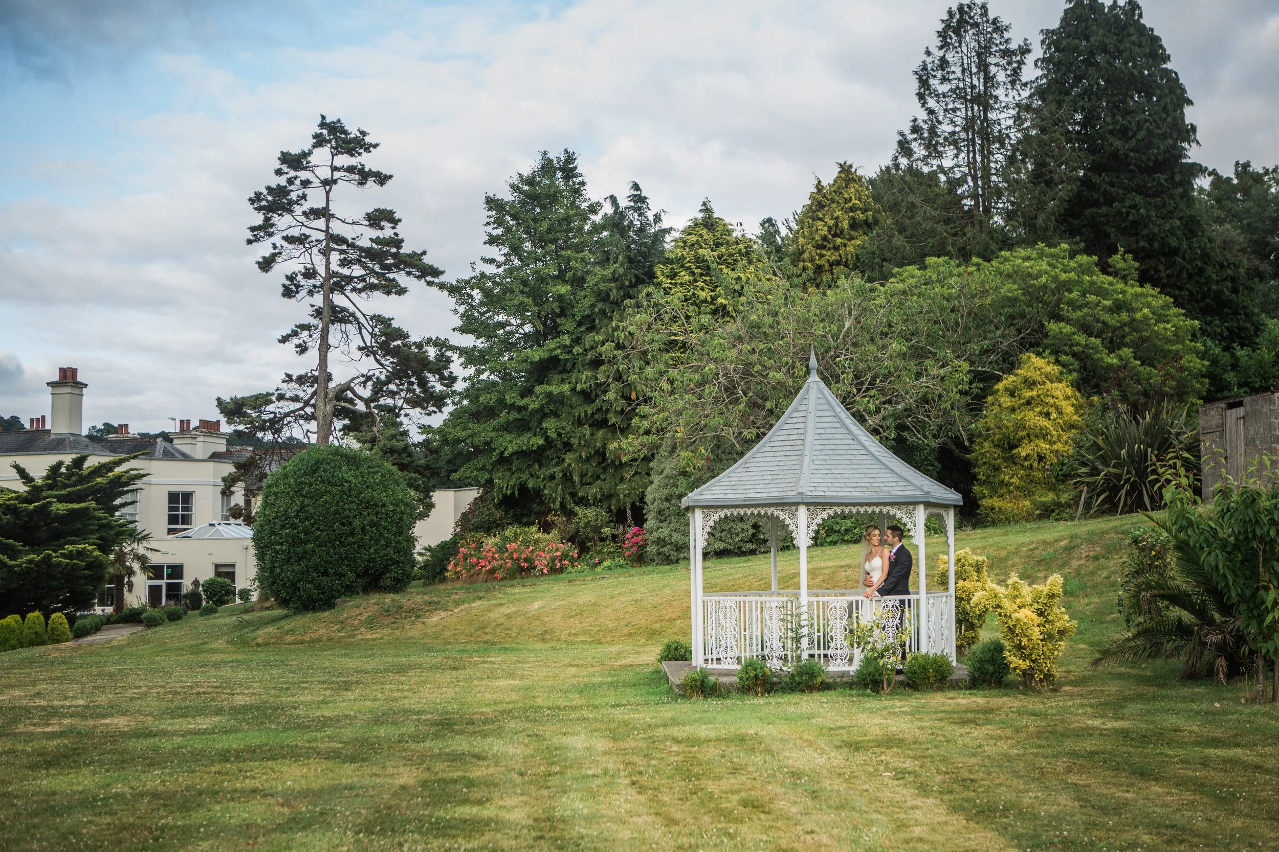 A bride and groom standing together inside a white gazebo on a grassy lawn, surrounded by trees and bushes, with a large house and a cloudy sky in the background.