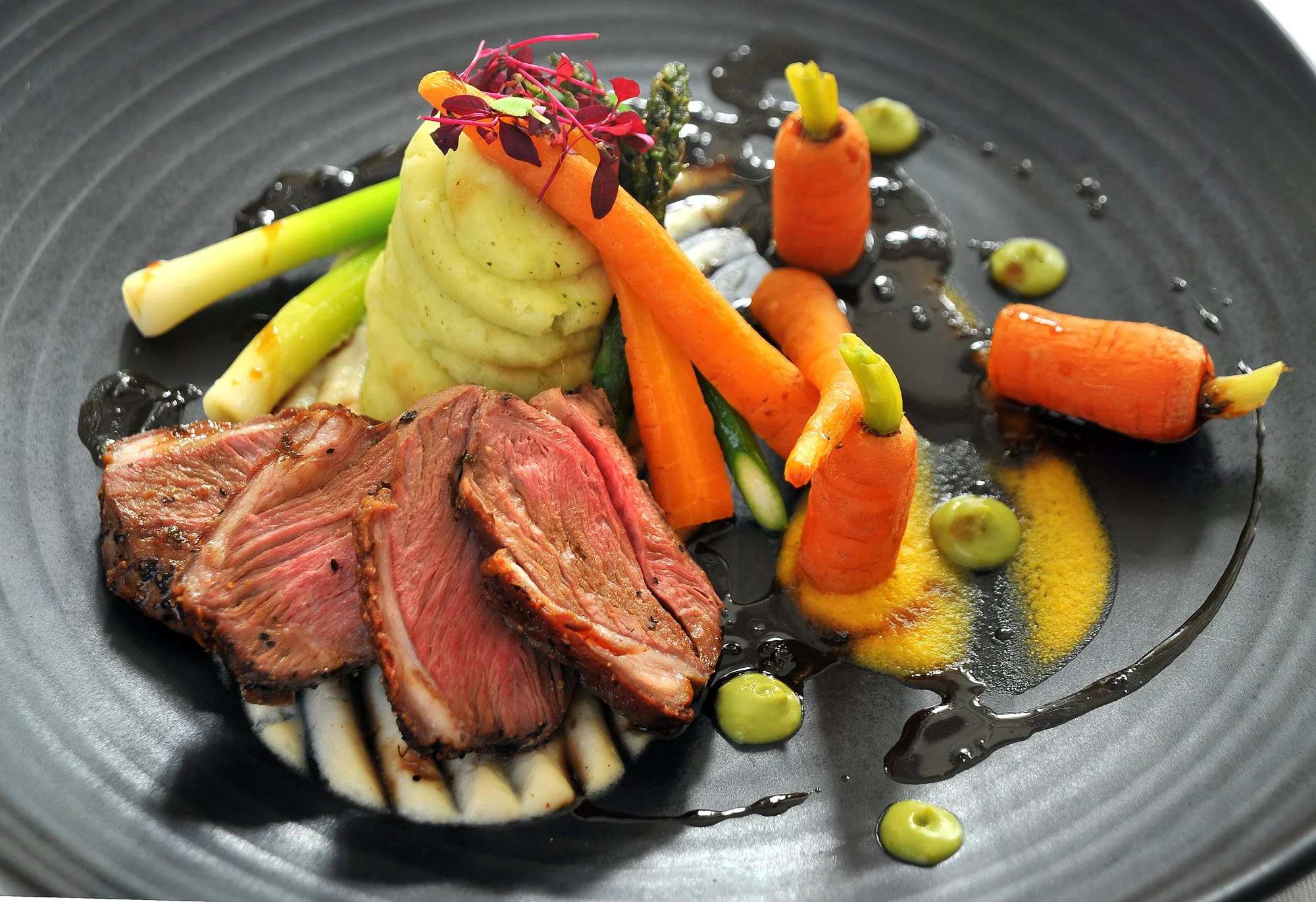 A gourmet dish featuring sliced medium-rare steak, mashed potatoes, and roasted baby carrots garnished with microgreens, drizzled with sauce, and accented with colorful vegetables on a black plate.
