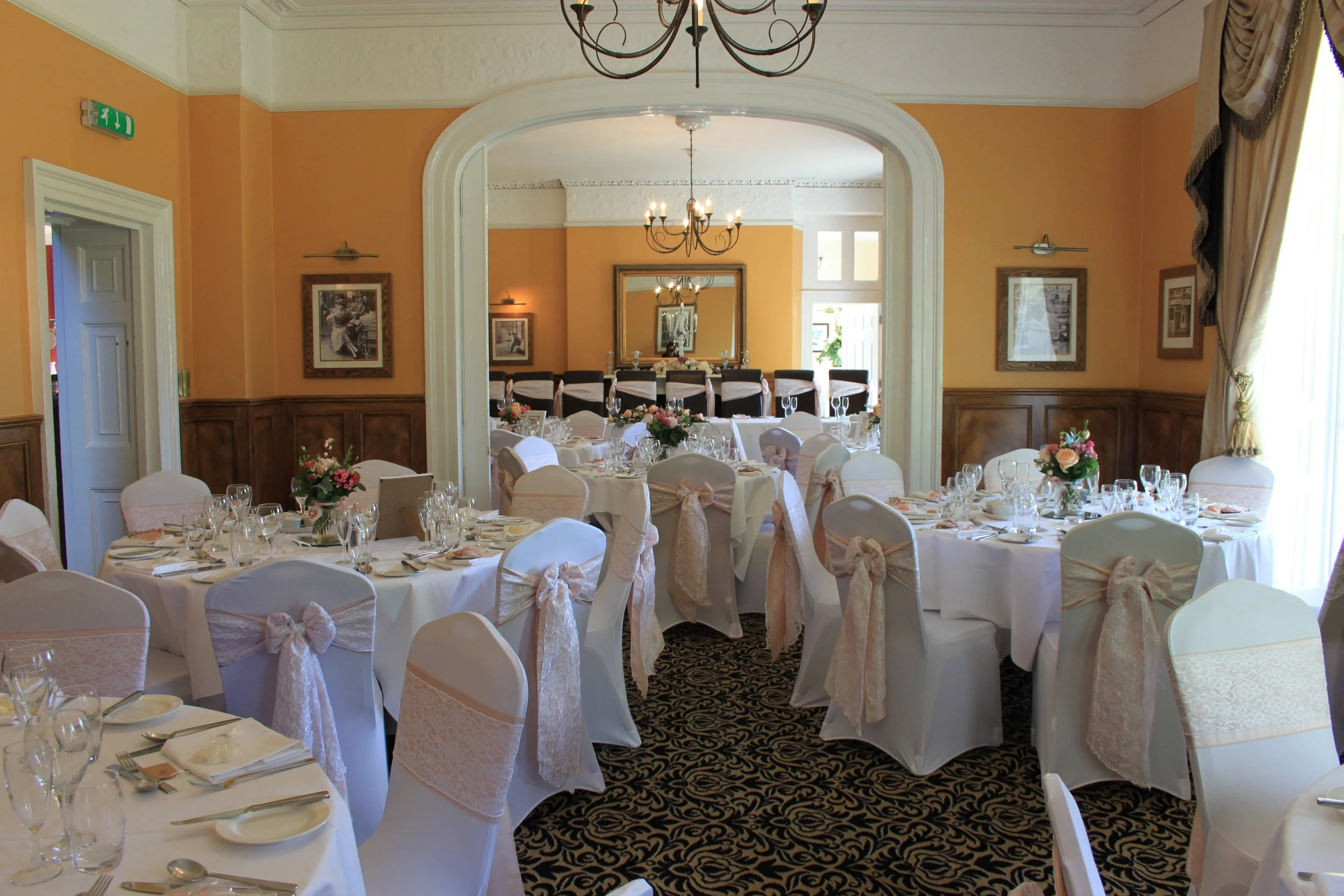 Wedding reception room decorated with round tables covered in white tablecloths, adorned with floral centerpieces, set with glassware, and chairs with lace covers and bows. There are chandeliers hanging from the ceiling, framed photographs on the wal