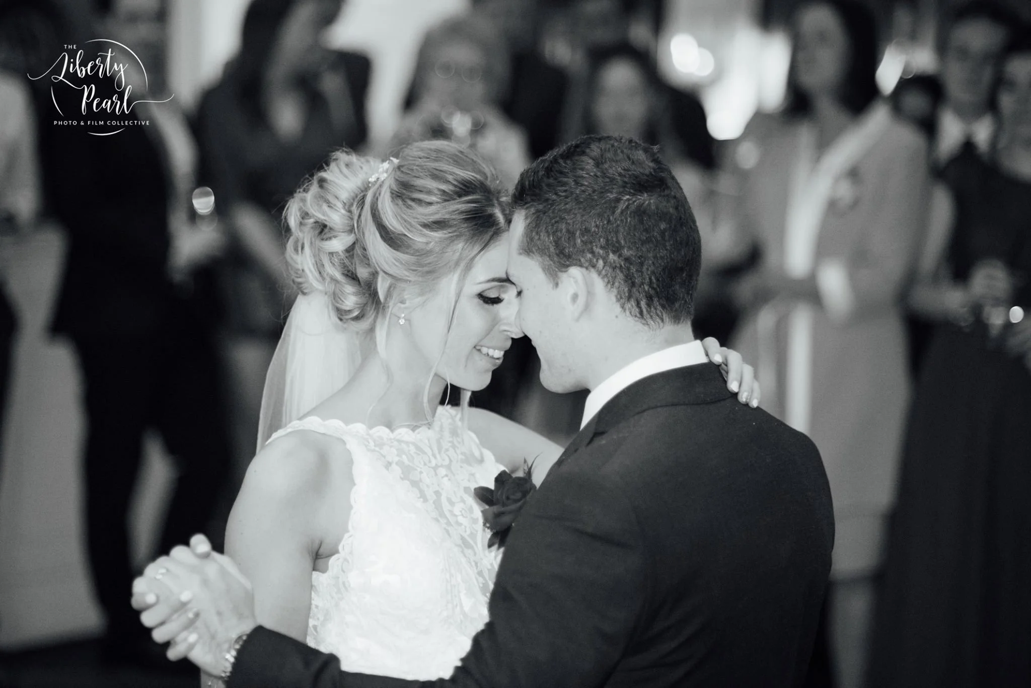 A bride and groom are dancing closely at their wedding reception, with their foreheads touching and eyes closed, surrounded by seated guests watching.