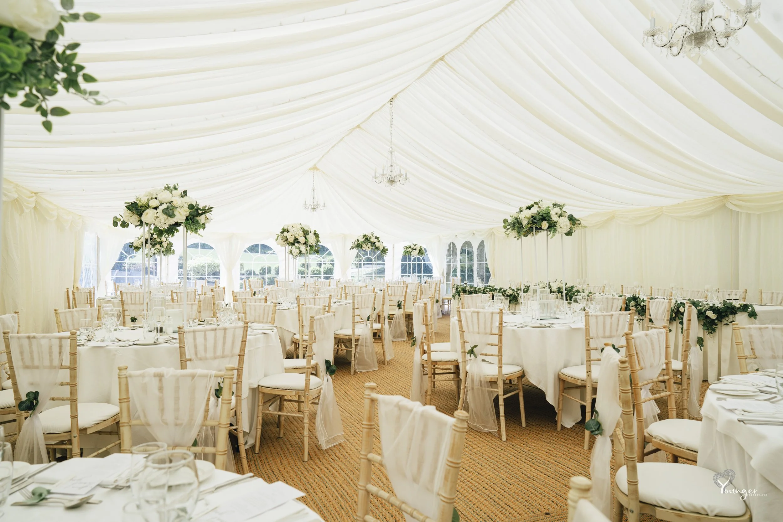 Elegant white wedding reception tent with round tables, floral centerpieces, and gold chairs, decorated with greenery and chandeliers.