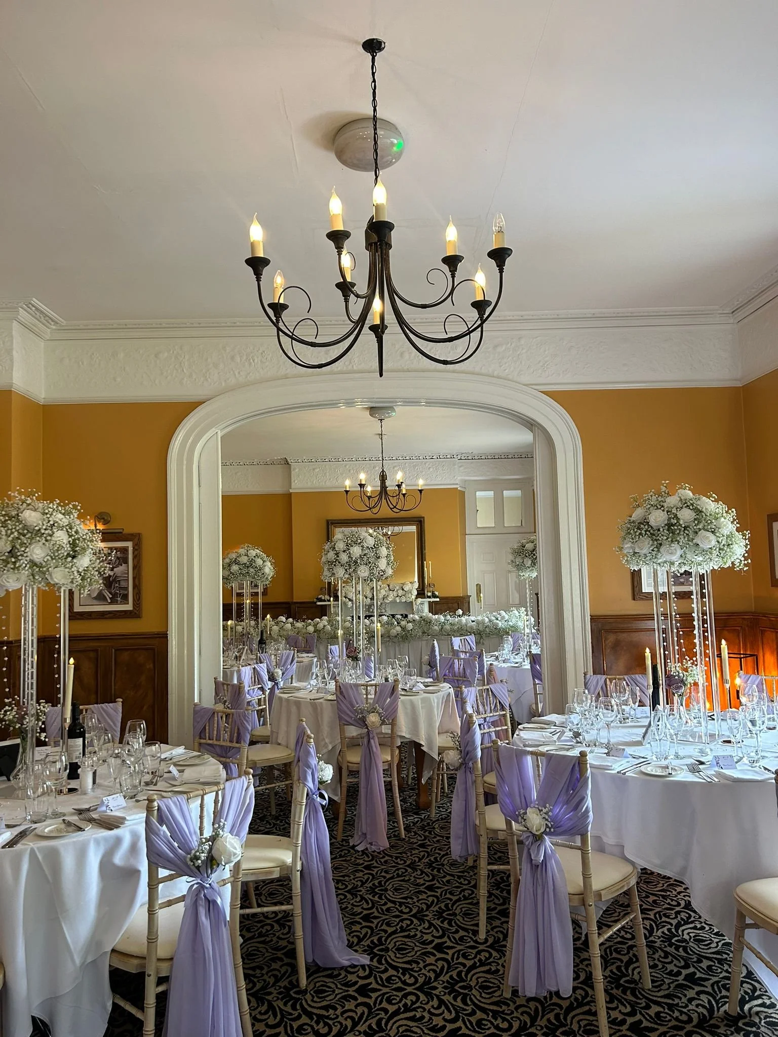 Elegant banquet hall decorated for a wedding or special event with round tables covered in white tablecloths, lavender sashes on chairs, and floral centerpieces. Chandeliers hang from the ceiling, and a mirror reflects the nicely decorated space.