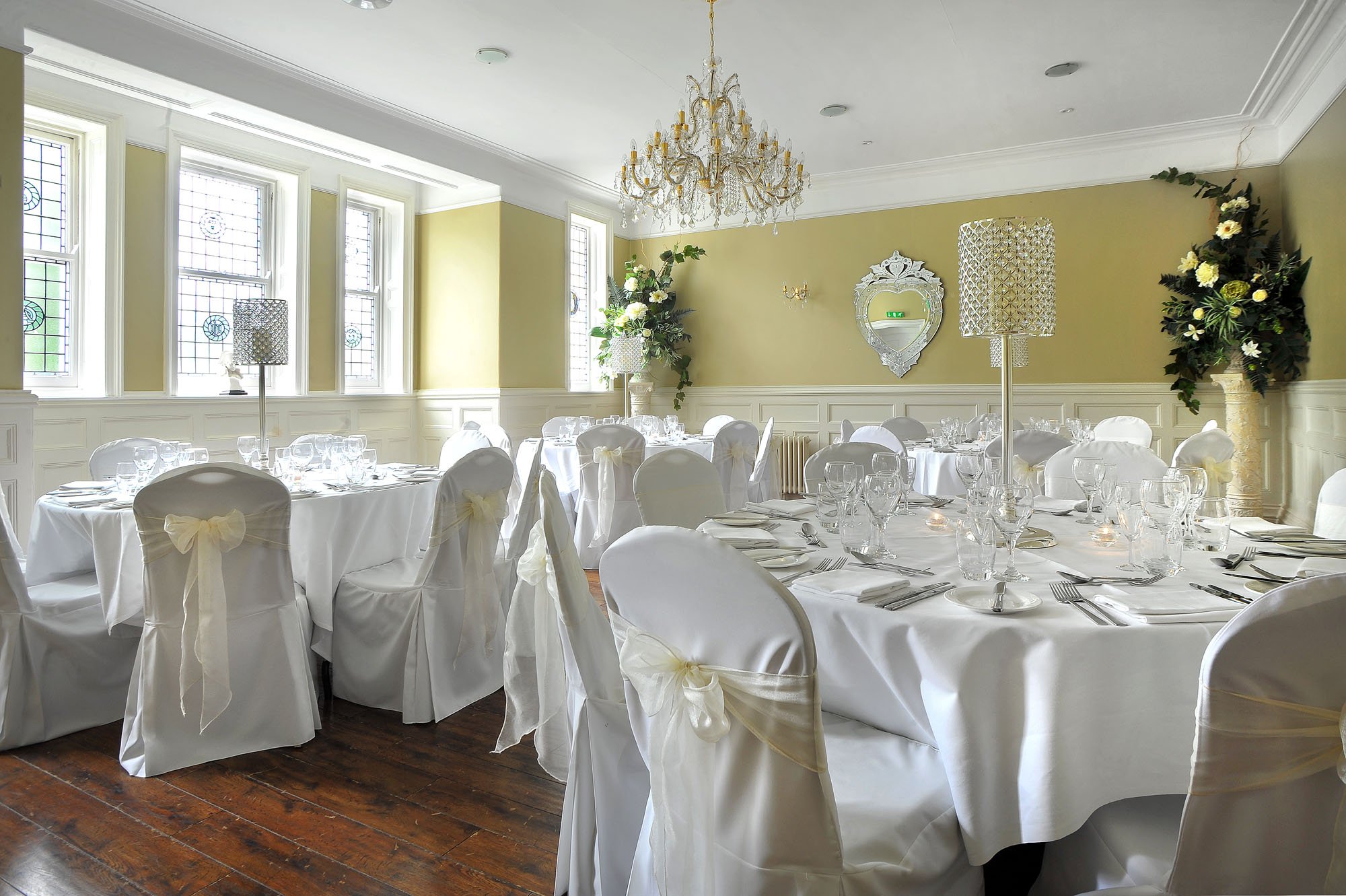 Elegant wedding reception setup with round tables covered in white tablecloths, fitted with white chairs adorned with bows, set with glassware, plates, and cutlery in a bright room with stained glass windows, floral arrangements, a chandelier, and decorative wall mirror.