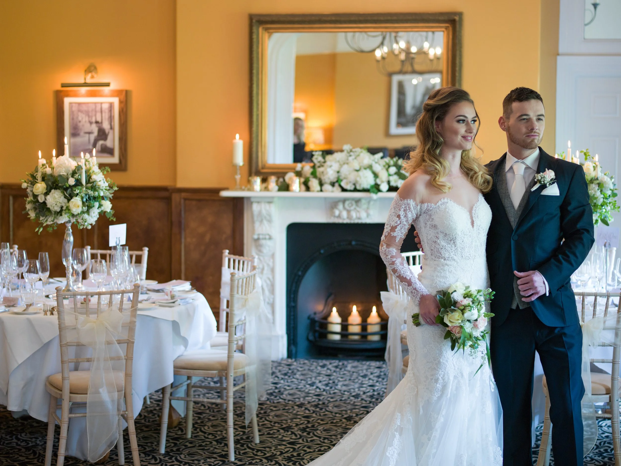 A bride and groom standing together at a wedding reception, with the bride holding a bouquet of flowers, in a decorated room with a fireplace, floral arrangements, and candles.