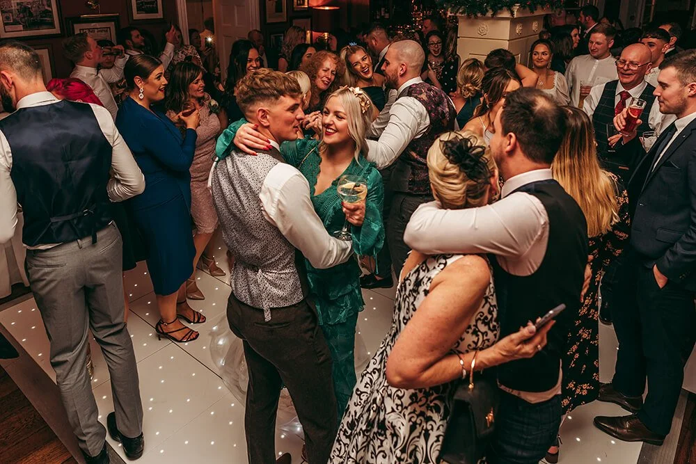 People socializing and dancing at a lively indoor party or celebration, with some holding drinks, in a warmly decorated room.