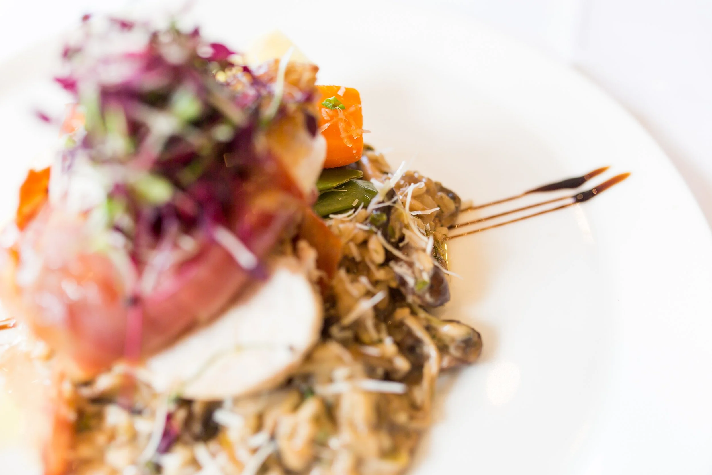 Close-up of a plated gourmet dish featuring a variety of ingredients, garnished with microgreens, with artistic sauce drizzles on a white plate.