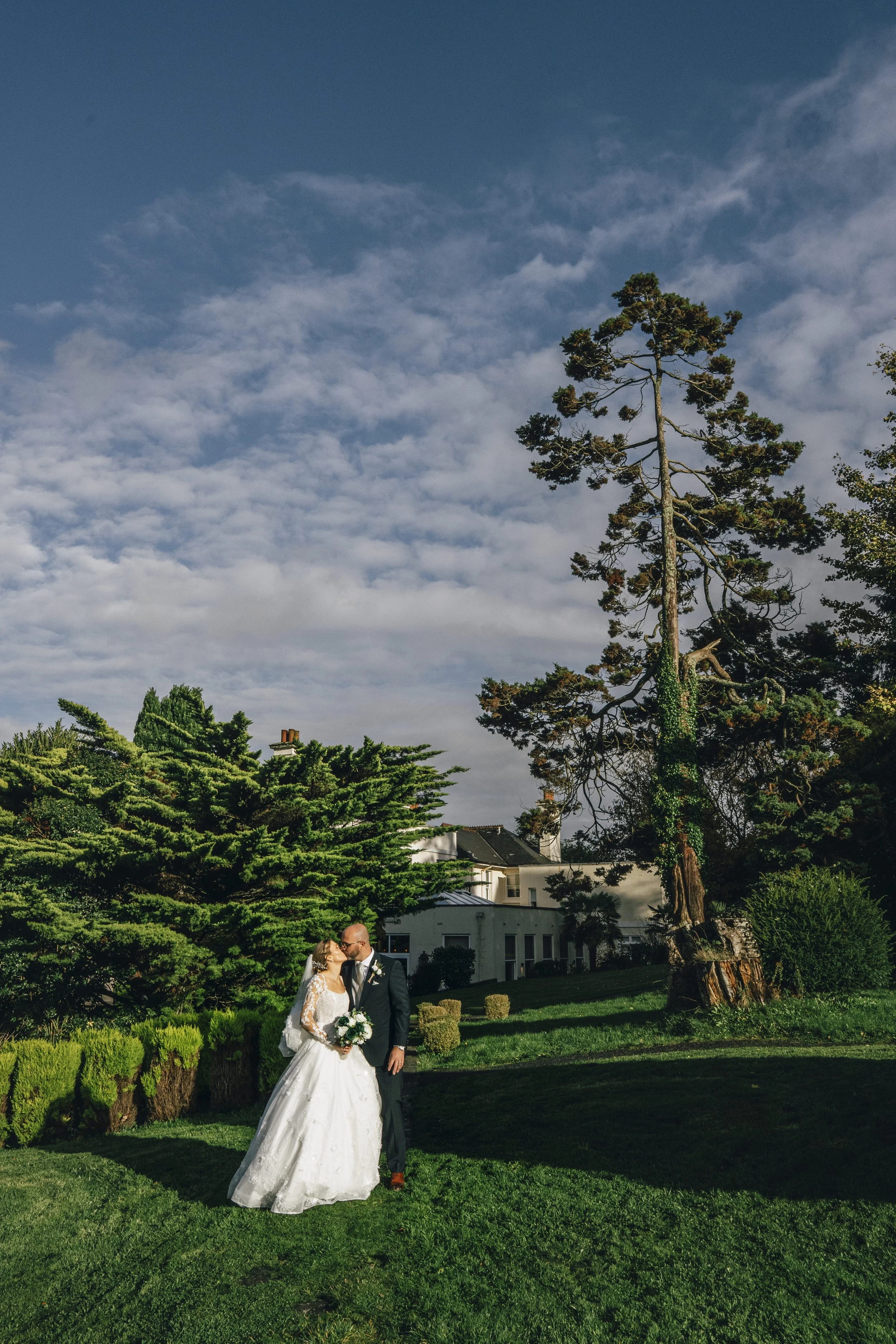 A bride and groom kissing outdoors in a garden with lush green trees and a large white house in the background.