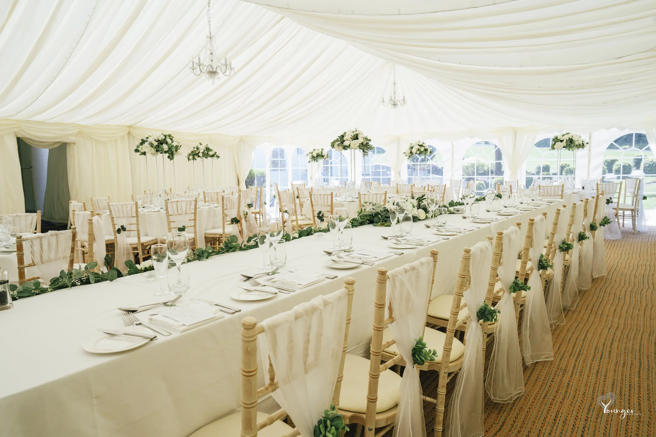 Elegant wedding reception setup in a large white tent with long dining tables decorated with white tablecloths, glassware, and floral centerpieces, surrounded by wooden chairs with white fabric and greenery embellishments, and chandeliers hanging fro