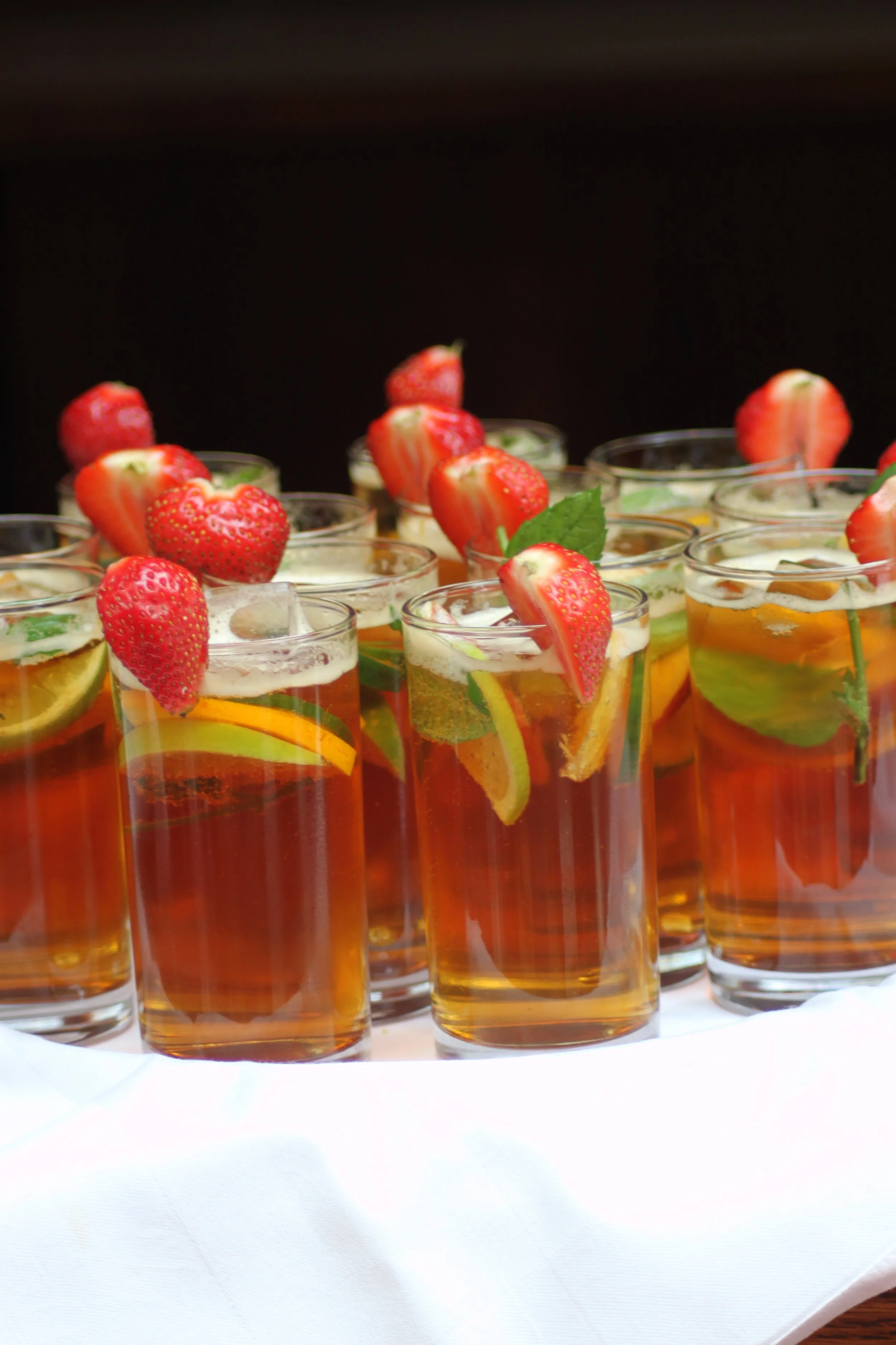 Glasses of iced tea garnished with strawberries, lemon slices, and mint leaves on a white tablecloth.