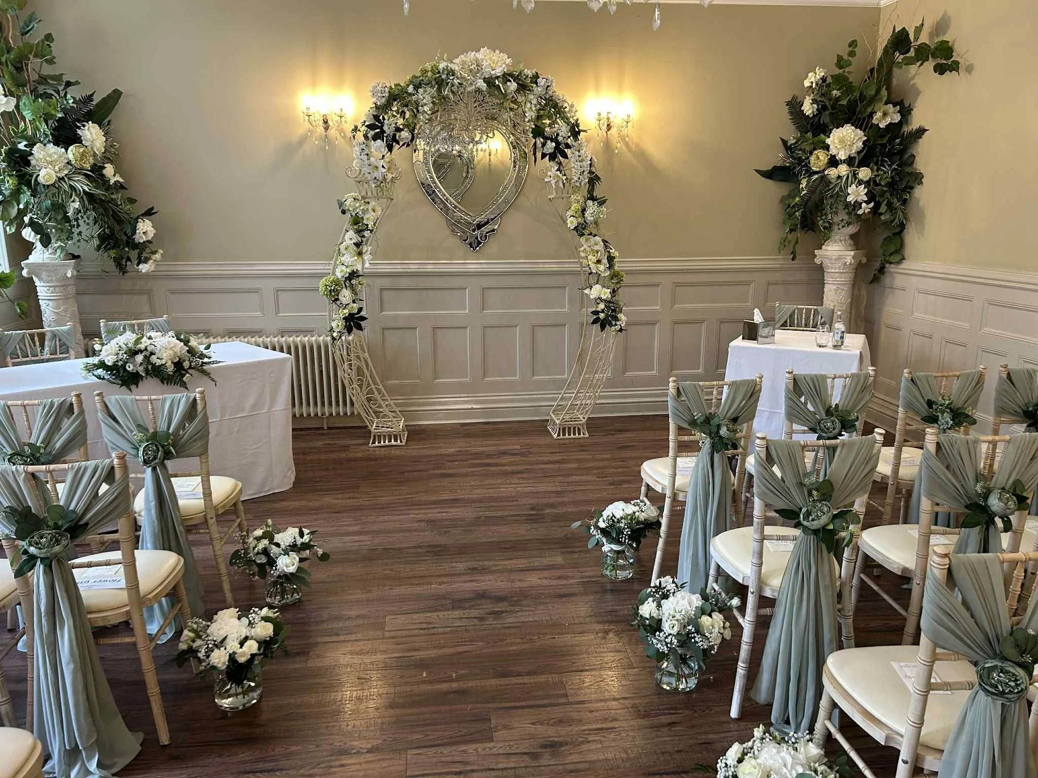 Indoor wedding ceremony setup with white chairs decorated with green and white fabric and flowers, floral arrangements on the floor, and a floral arch at the altar with a heart-shaped mirror and floral decorations.
