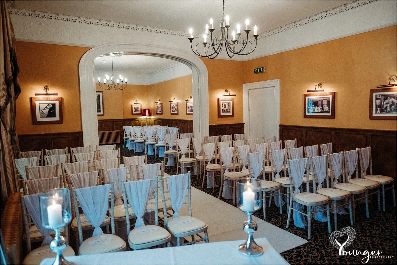 Empty wedding or event ceremony room with rows of white chairs draped with fabric and candles on the tables, warmly lit with chandeliers and wall lights.