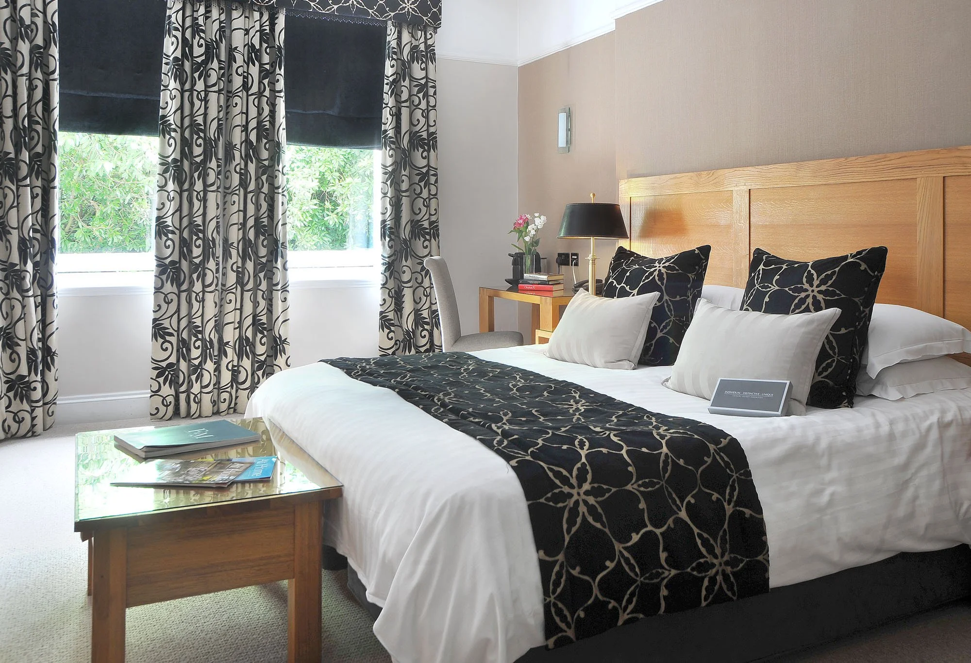 A neatly made hotel bed with black and white patterned pillows and a matching bed runner, a wooden headboard, a bedside table with a black lamp, a vase of flowers, and a chair next to two windows with black and white patterned curtains.