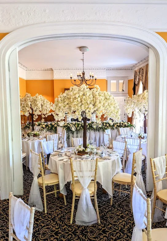 Elegant banquet hall with round tables decorated with white tablecloths, floral centerpieces with white flowers, surrounded by gold chairs with white sashes, ornate orange and white walls, and a chandelier hanging from the ceiling.