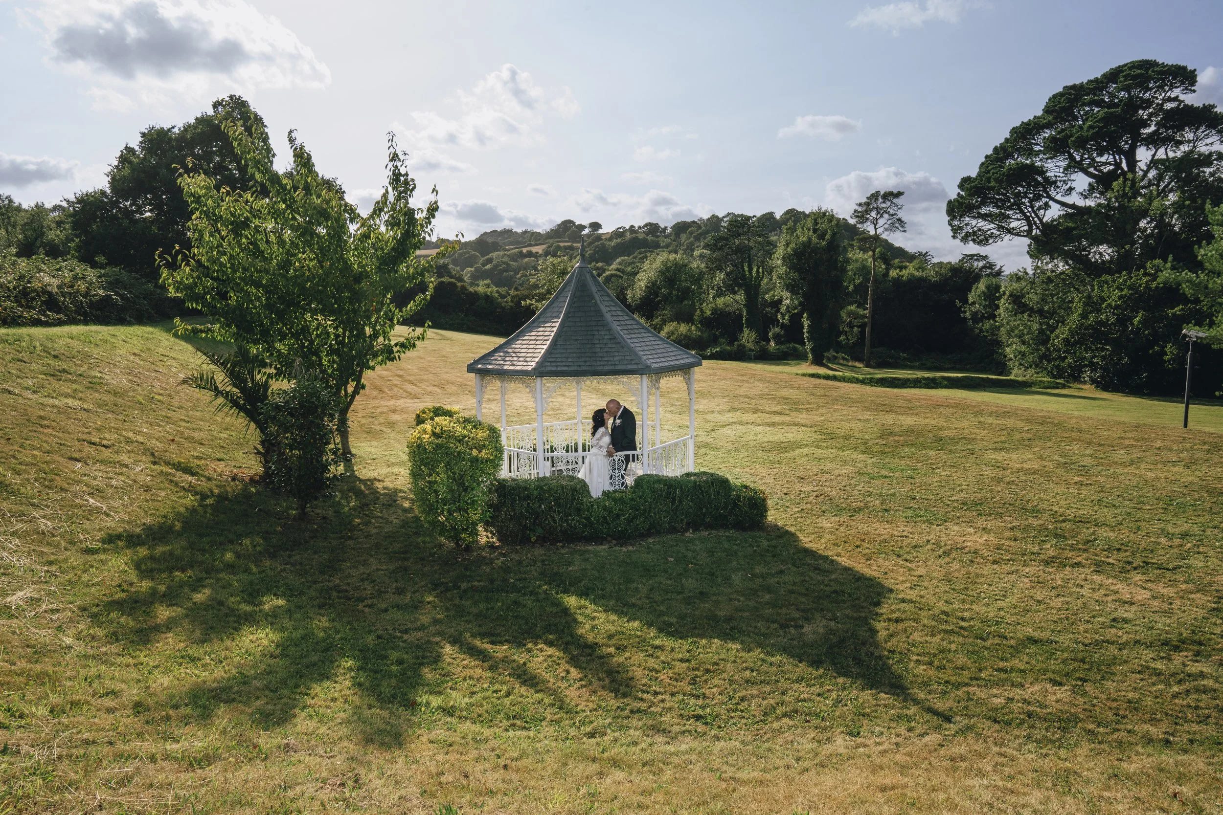 A couple dressed in wedding attire standing inside a white gazebo in a grassy park, surrounded by trees under a partly cloudy sky.