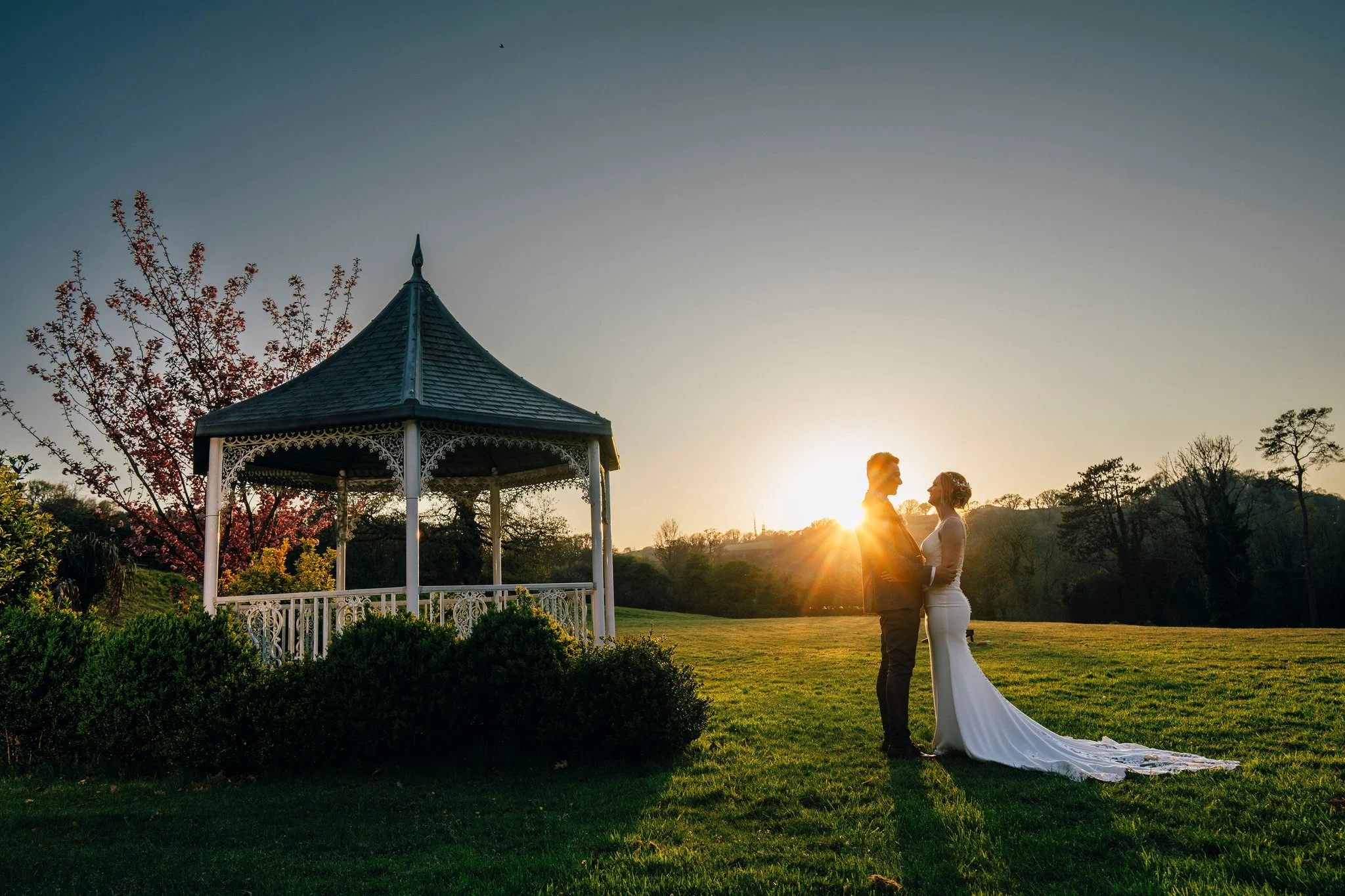 A bride and groom standing close together at sunset in a grassy field, facing each other, with a wedding gown train trailing on the ground. A gazebo is visible to the left, with trees in the background.