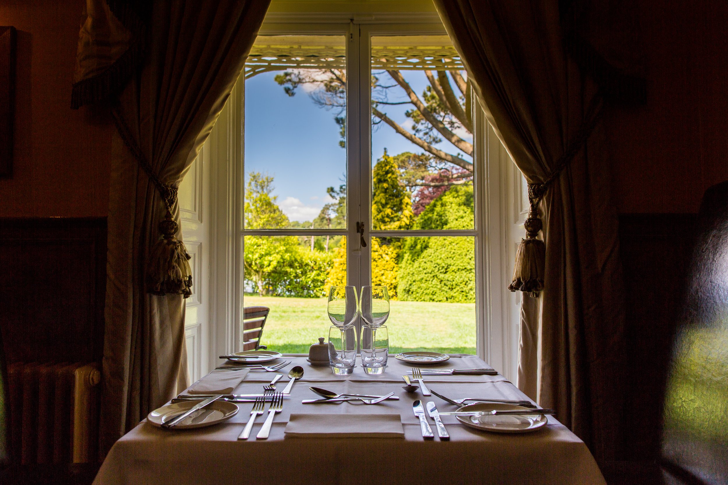 A dining table set for four with plates, glasses, and silverware, positioned in front of a large window showing a lush green garden and trees outside under a blue sky.
