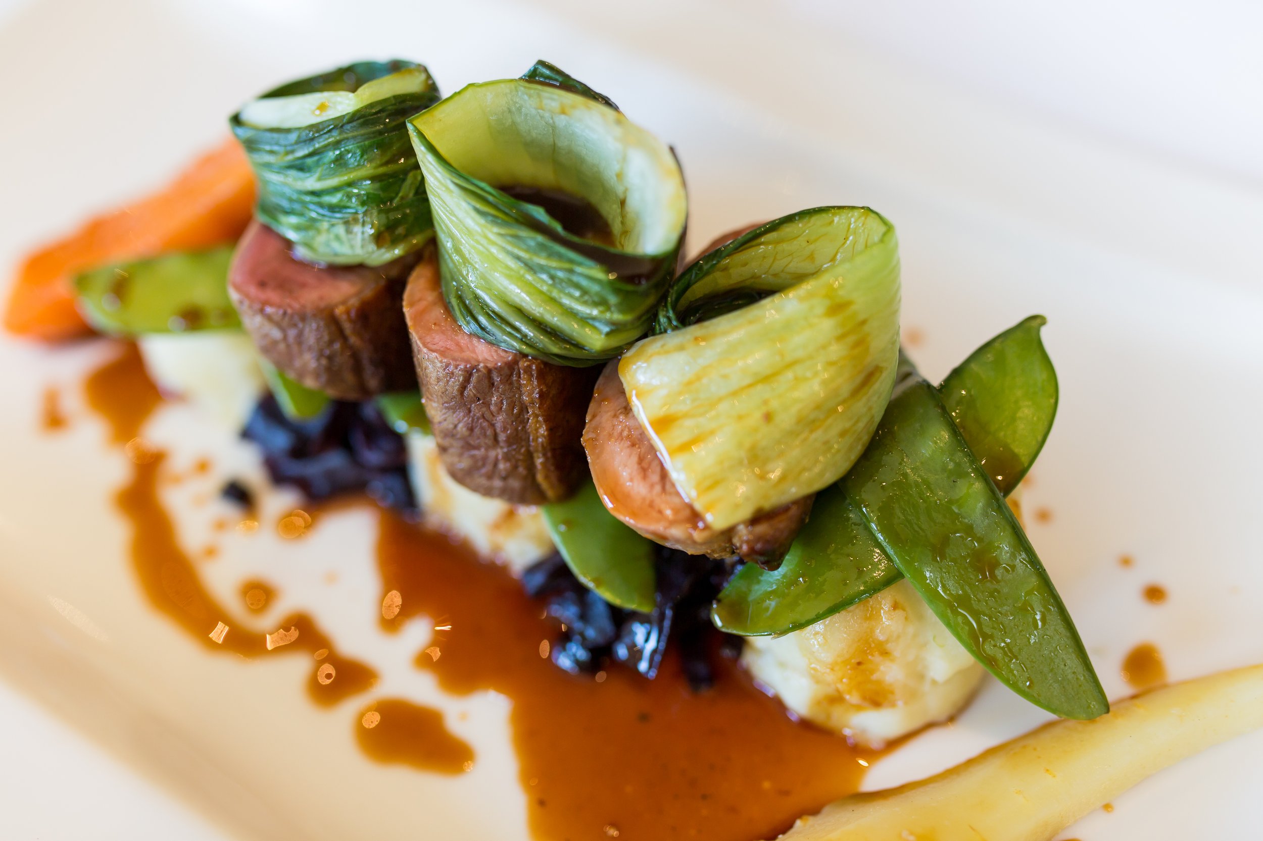 Gourmet plated dish with slices of seared meat, roasted vegetables, and green garnishes, topped with a brown sauce.