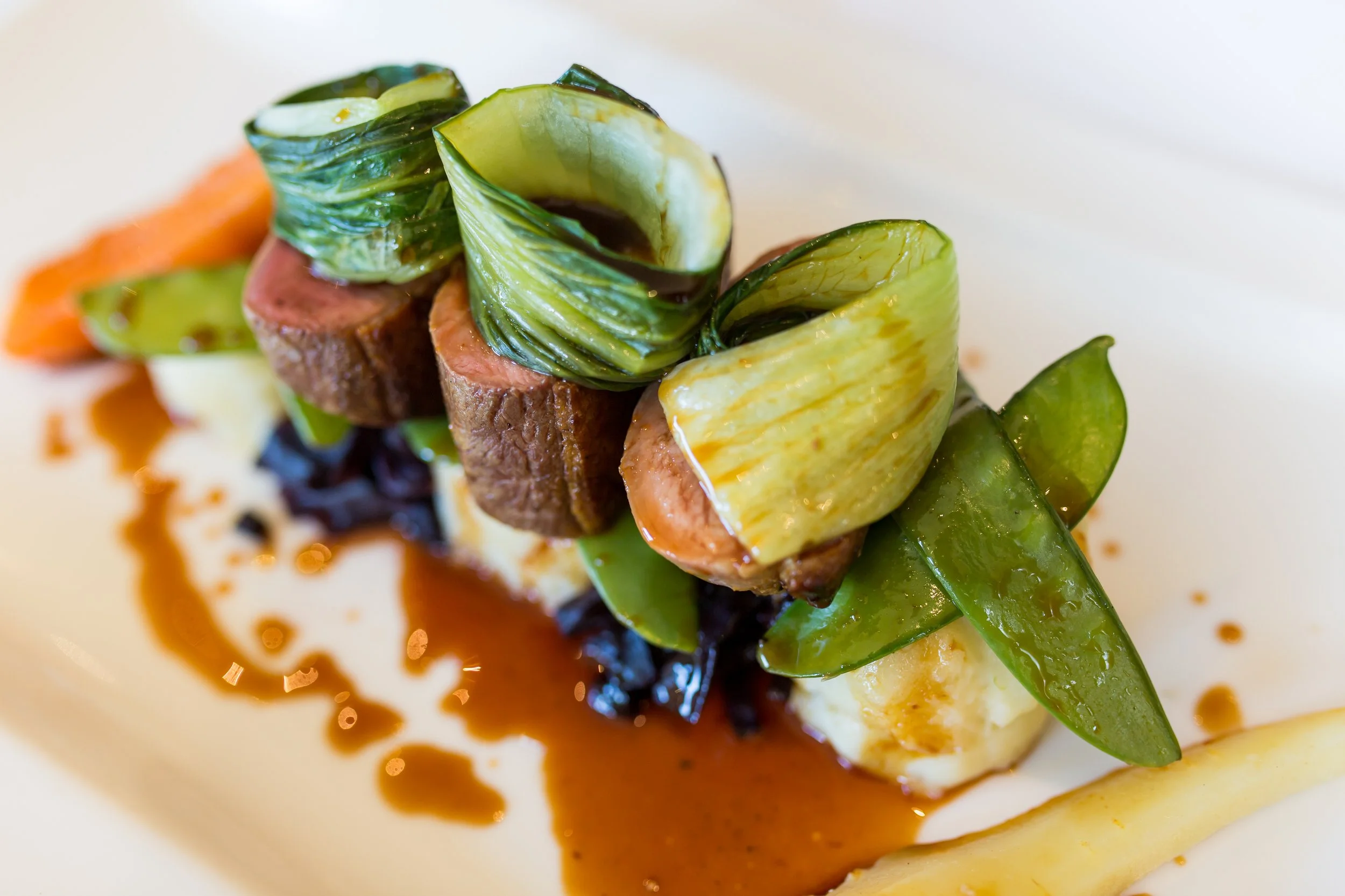 A gourmet dish featuring slices of cooked meat topped with green vegetable curls, served with various vegetables and drizzled with brown sauce.