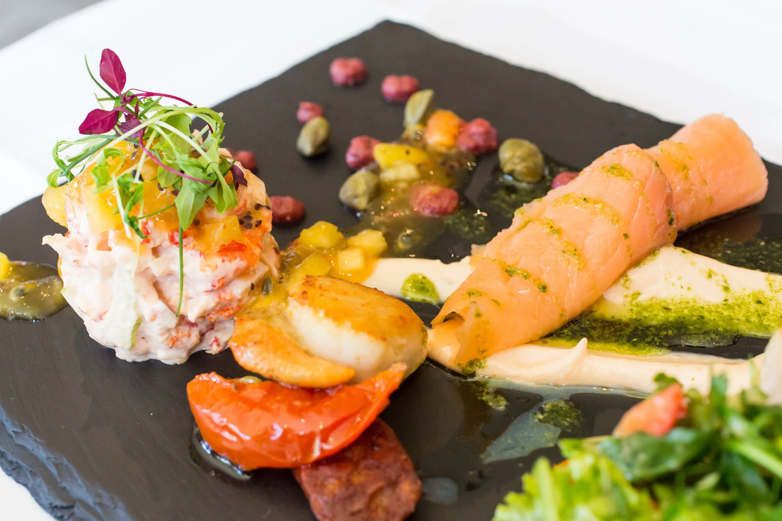 Gourmet dish with a scoop of potato salad topped with microgreens, presented with roasted cherry tomatoes, a slice of smoked salmon with green sauce, and garnished with colorful berries and herbs, served on a black slate plate.