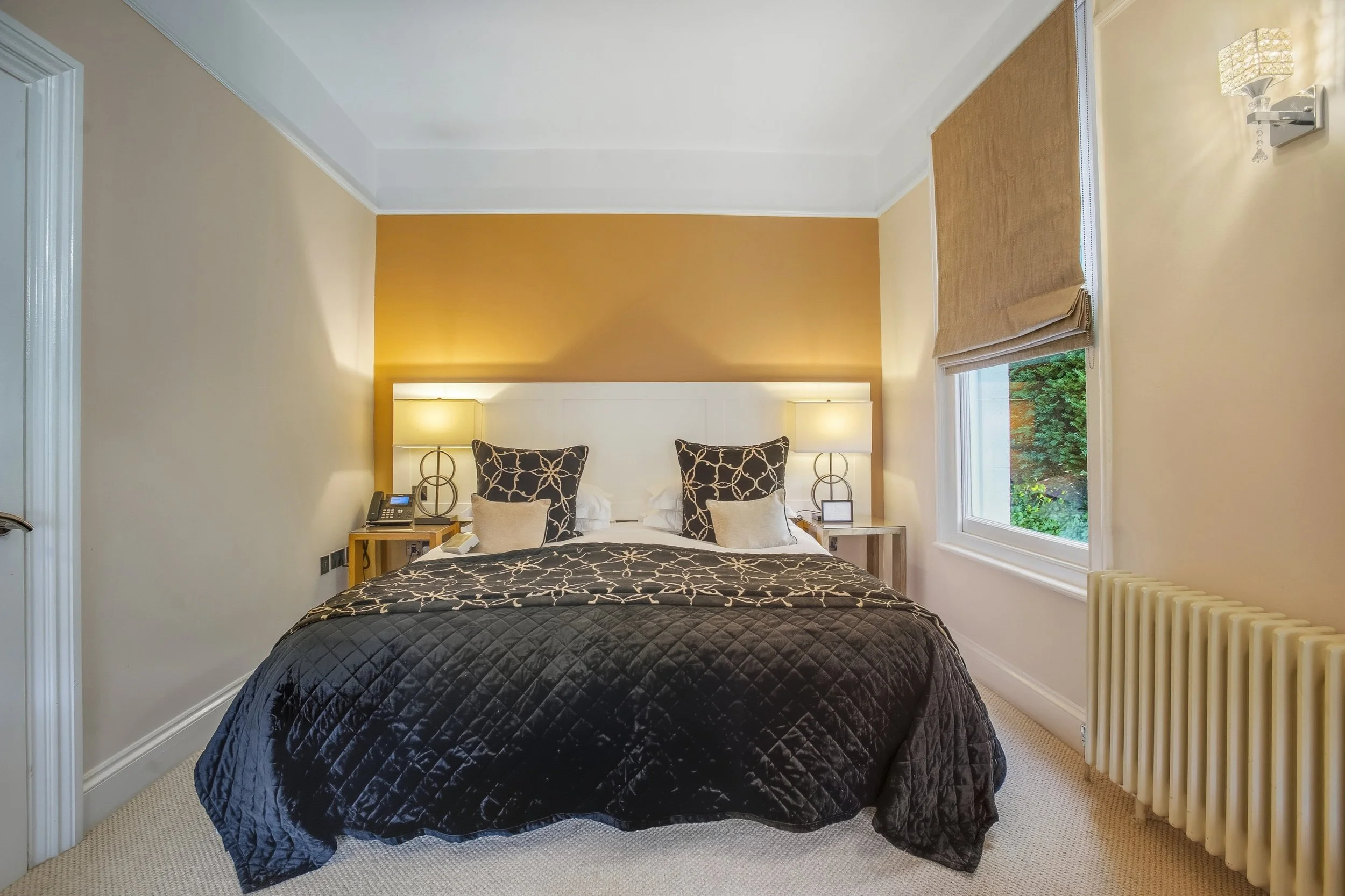 A neatly made bed with black and beige patterned pillows and a black quilt, flanked by two wooden nightstands with lamps, in a hotel room with a yellow accent wall and a window with a beige roman shade.
