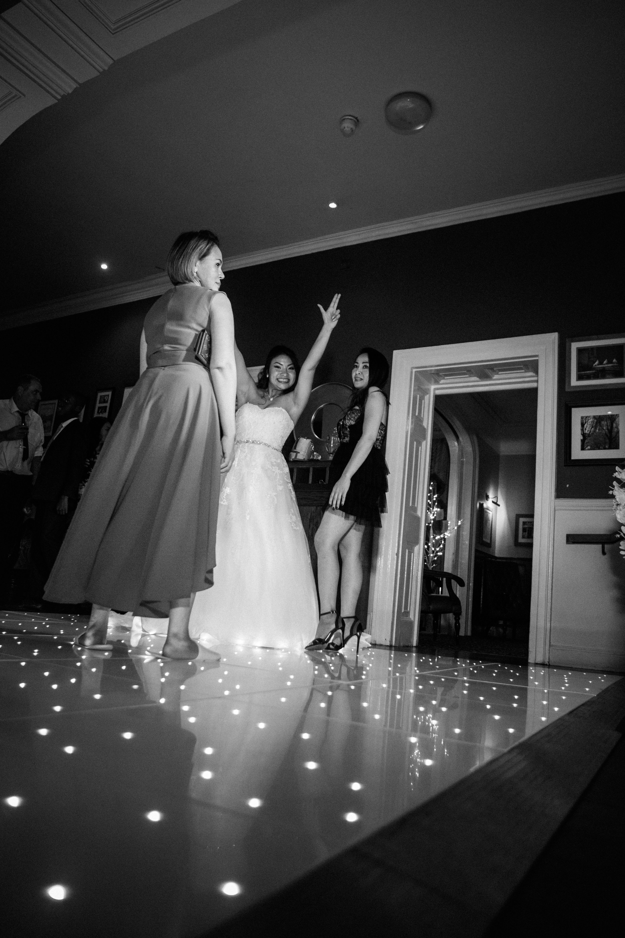 Black and white photo of three women dancing at a wedding reception, with a bride in a wedding dress and two women in dresses, one in a black dress with high heels.