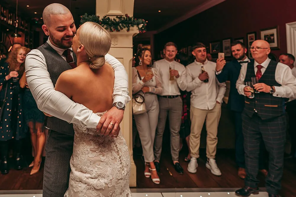 A wedding reception with a bride and groom dancing closely, surrounded by guests watching and taking photos in a warmly decorated room.