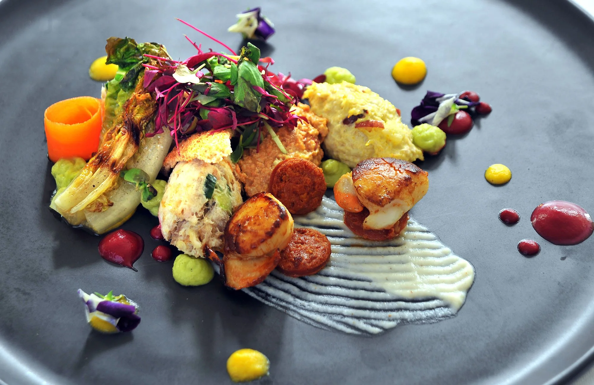An artfully plated gourmet dish with seared scallops, vegetables, microgreens, and colorful sauces on a dark plate.
