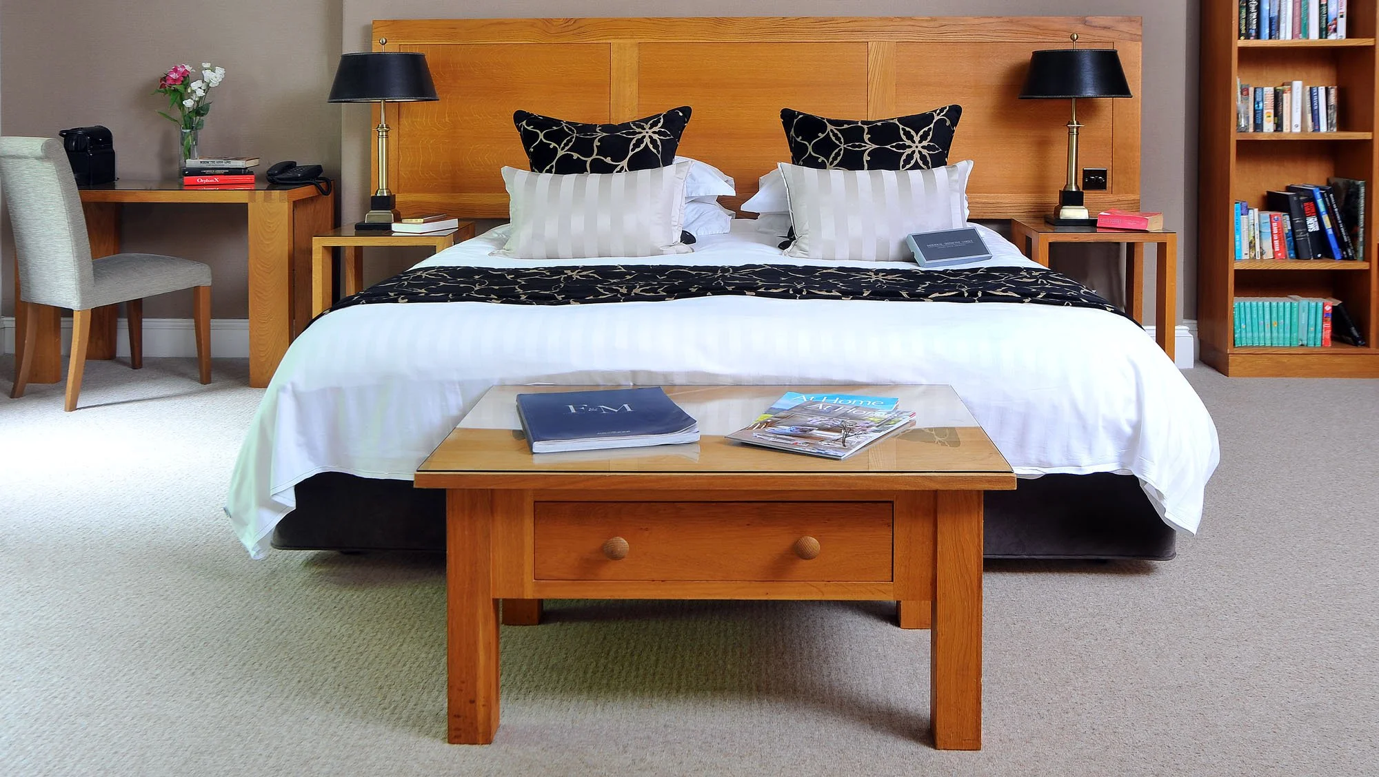 A neatly made hotel bed with white linens, black and gold pillows, and a black and gold bed runner, flanked by wooden nightstands with black lamps. A wooden coffee table with magazines and a blue folder is at the foot of the bed. To the left is a wooden desk with a chair, flowers, and a telephone. To the right, a wooden bookshelf filled with books is visible.