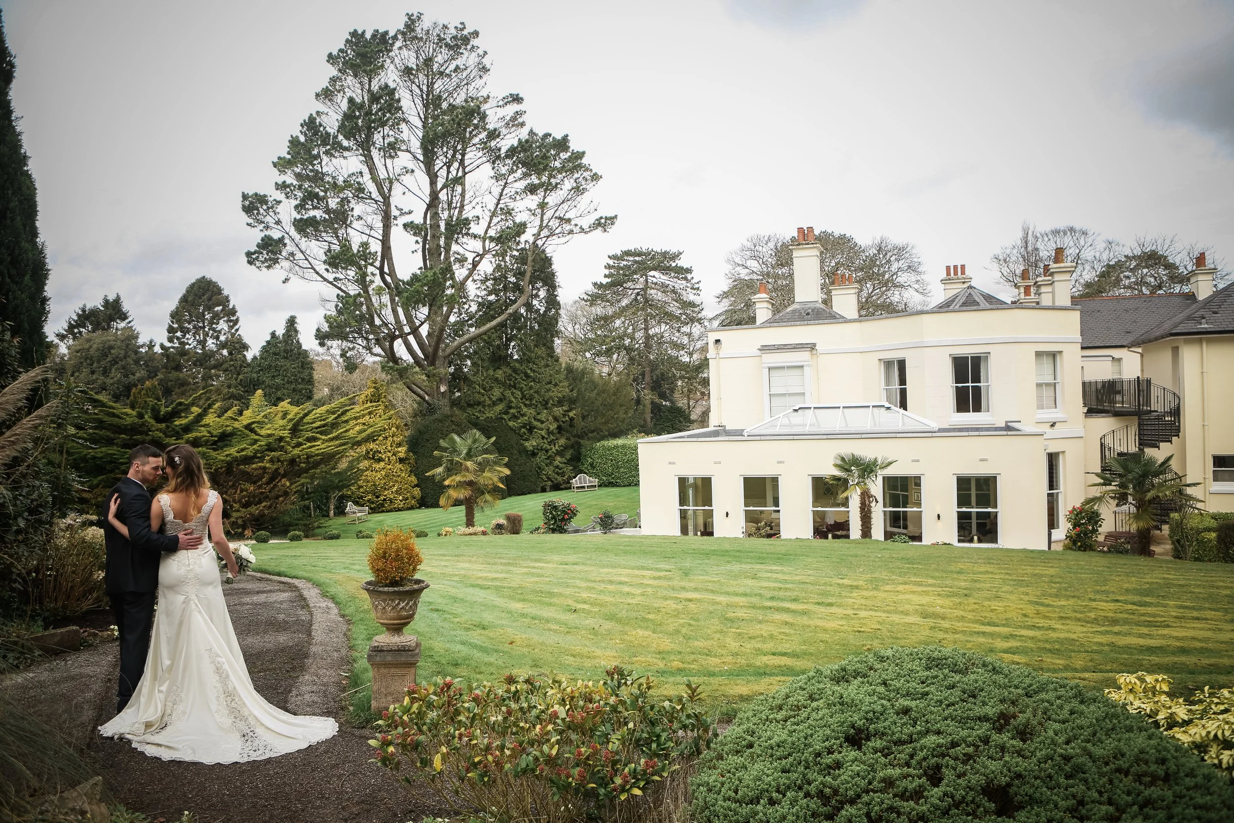 A bride and groom standing close together on a garden path, with a large white house and lush greenery in the background.