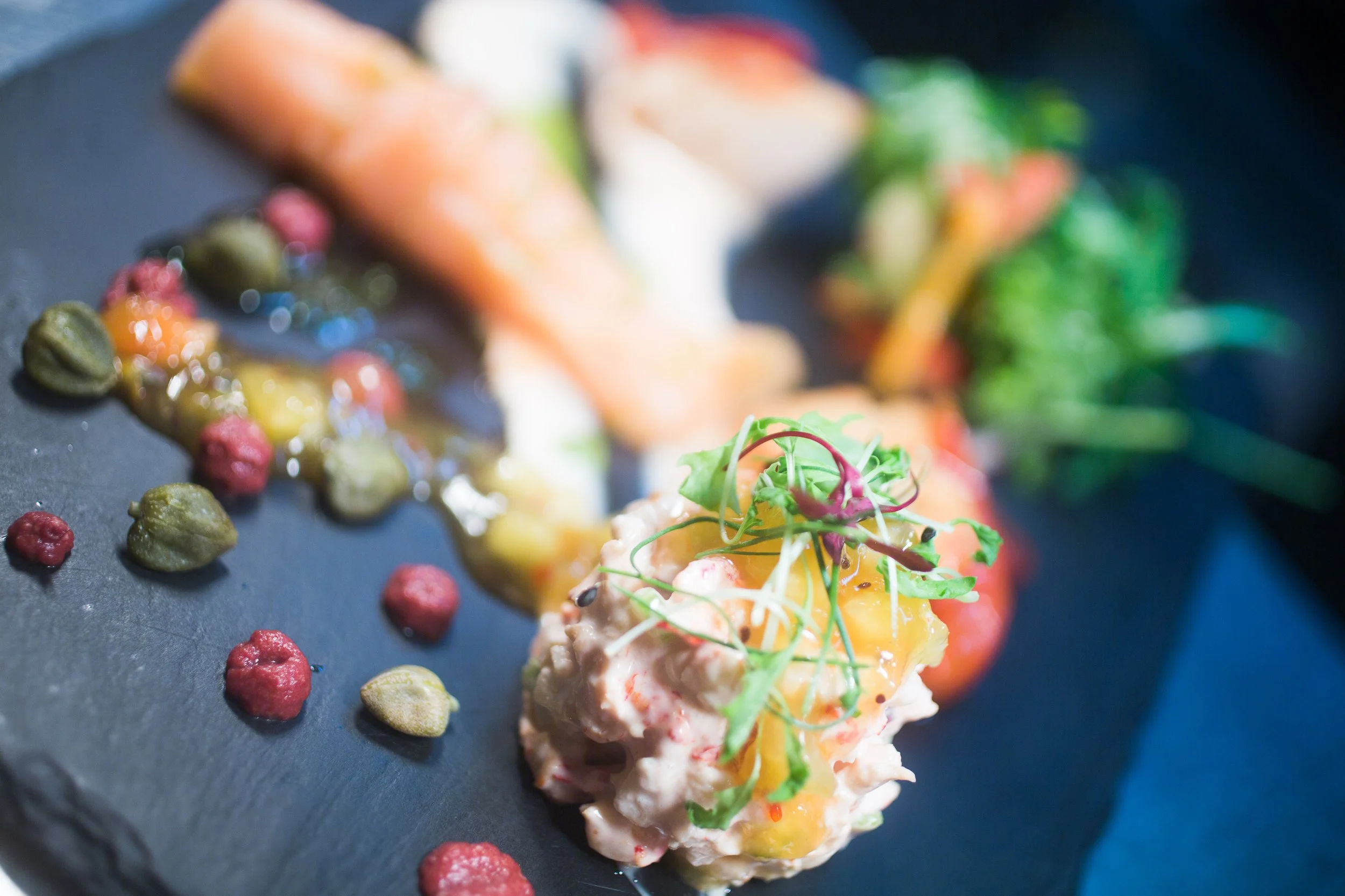 Gourmet dish with smoked salmon, a dollop of potato salad topped with an egg yolk, microgreens, and scattered pink and green peppercorns on a black slate plate.