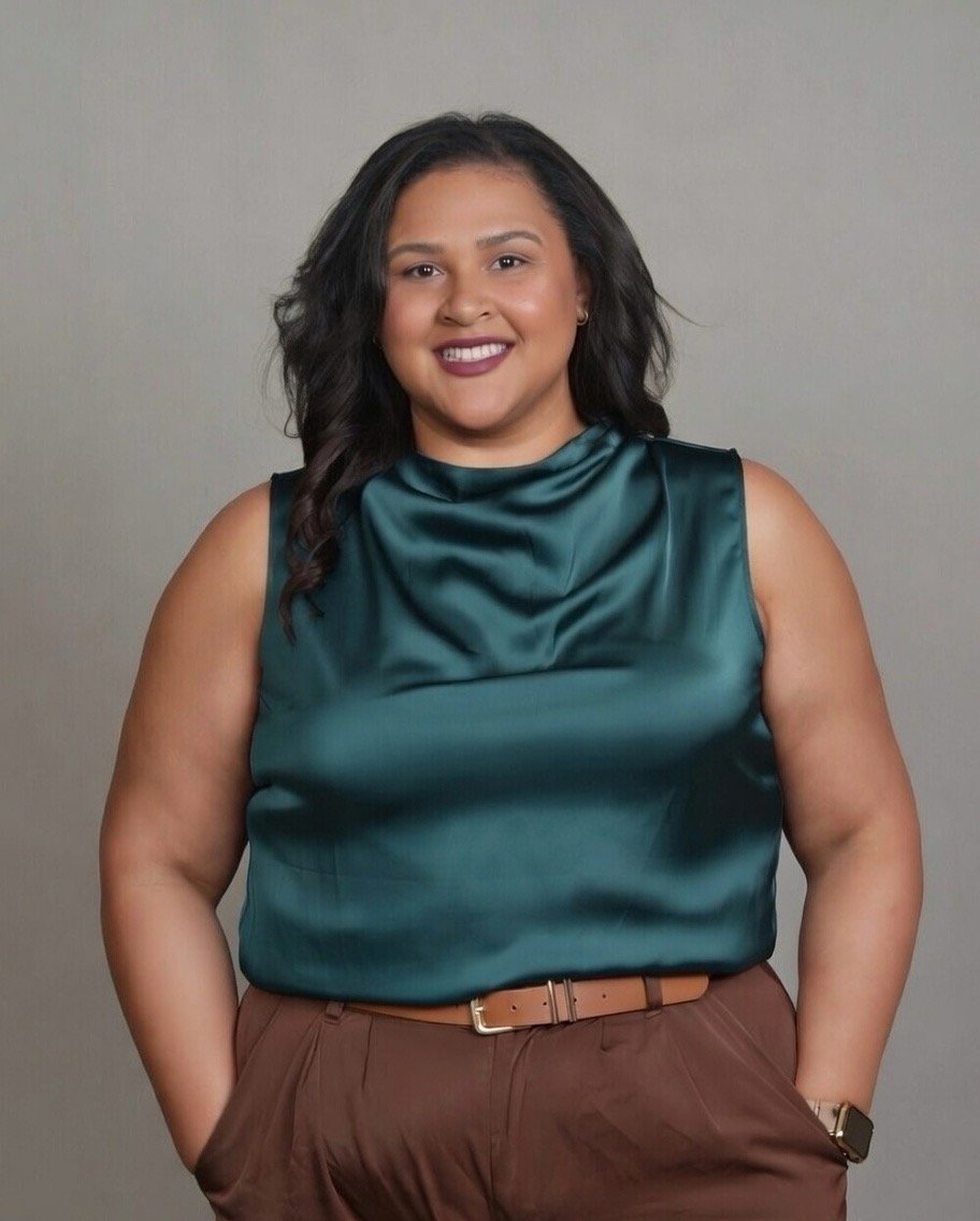 A woman with dark hair and medium skin, wearing a teal sleeveless satin blouse and brown pants with a belt, smiling against a plain background.