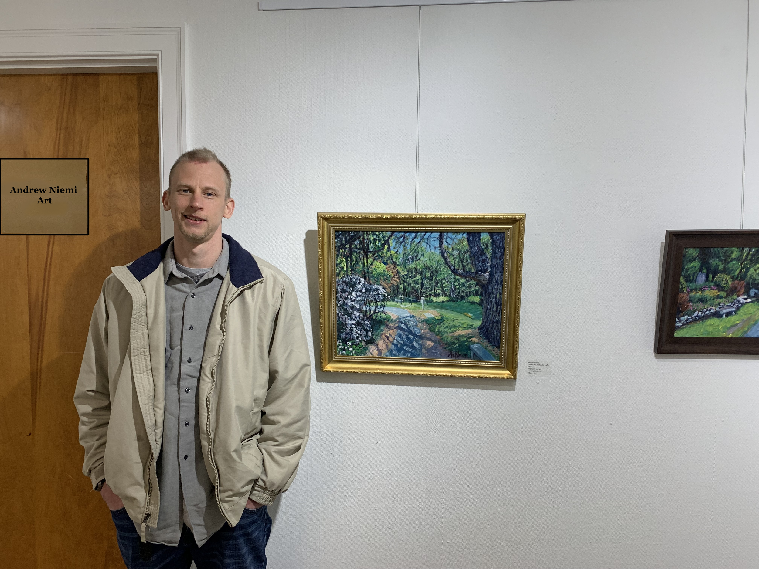 A young man with light hair and a light beard standing in an art gallery near framed paintings of outdoor landscapes. He is wearing a beige jacket over a gray shirt and has his hands in his pockets.
