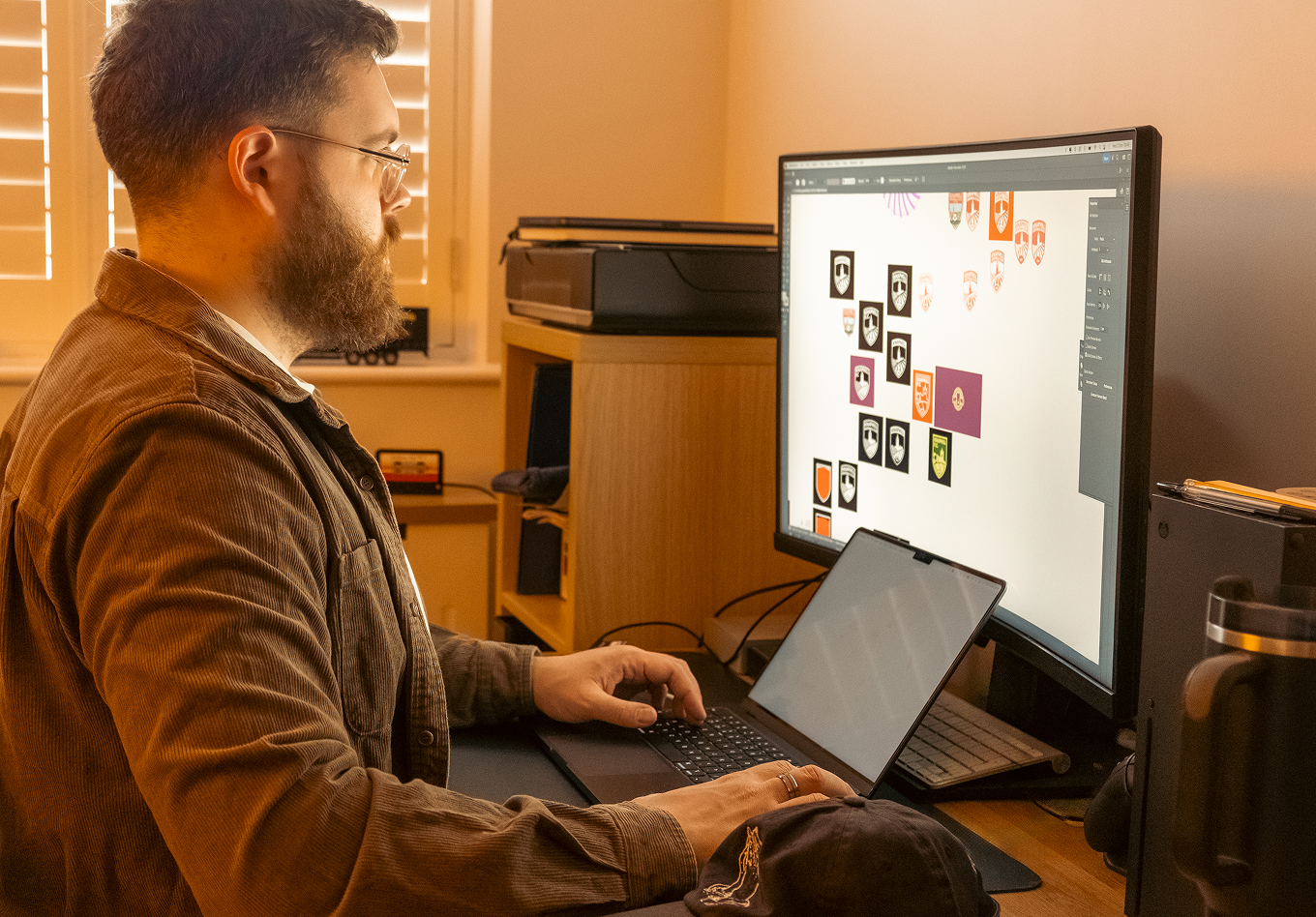 A man with glasses and a beard working on a digital design of crest logos on a large computer monitor with a tablet and keyboard in front of him in a home office.