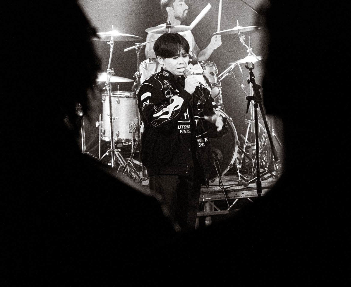 Black and white photo of a young man singing into a microphone on stage with a band in the background, seen through a hole or frame, with silhouettes of people in the foreground.