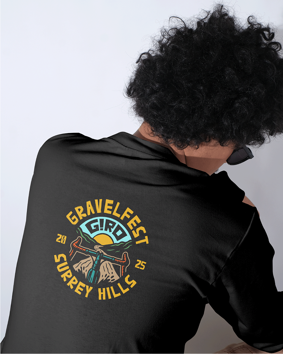 Person with curly hair wearing a black shirt with a colorful 'Travelfest Giron Jersey Hills' logo on the back, showing a mountain, a waterfall, and a sunrise or sunset background.