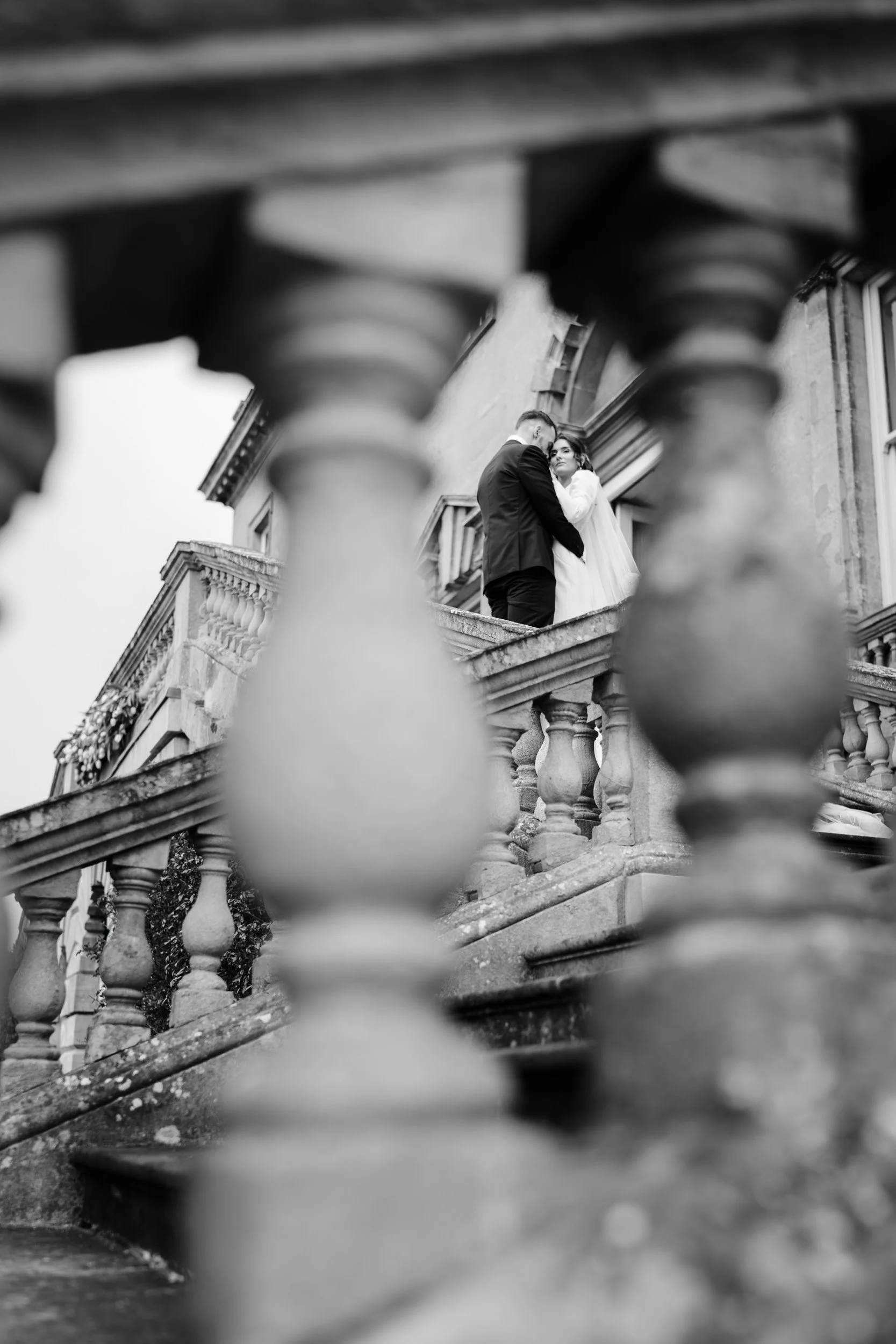 Bride and groom embracing on the balcony at Kirtlington Park in Oxfordshire, photographed through the stone balustrode