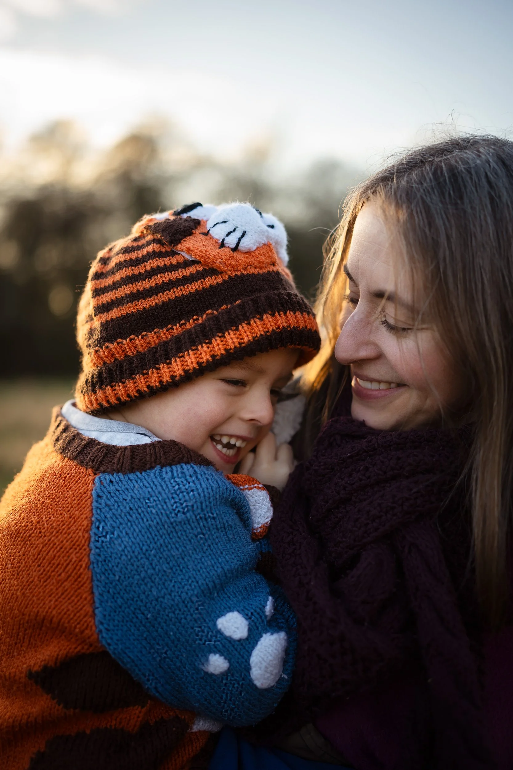 A mother smiles at her happy boy wearing a bobble hat in a relaxed outdoor family photography session in Amersham, Buckinghamshire a golden hour
