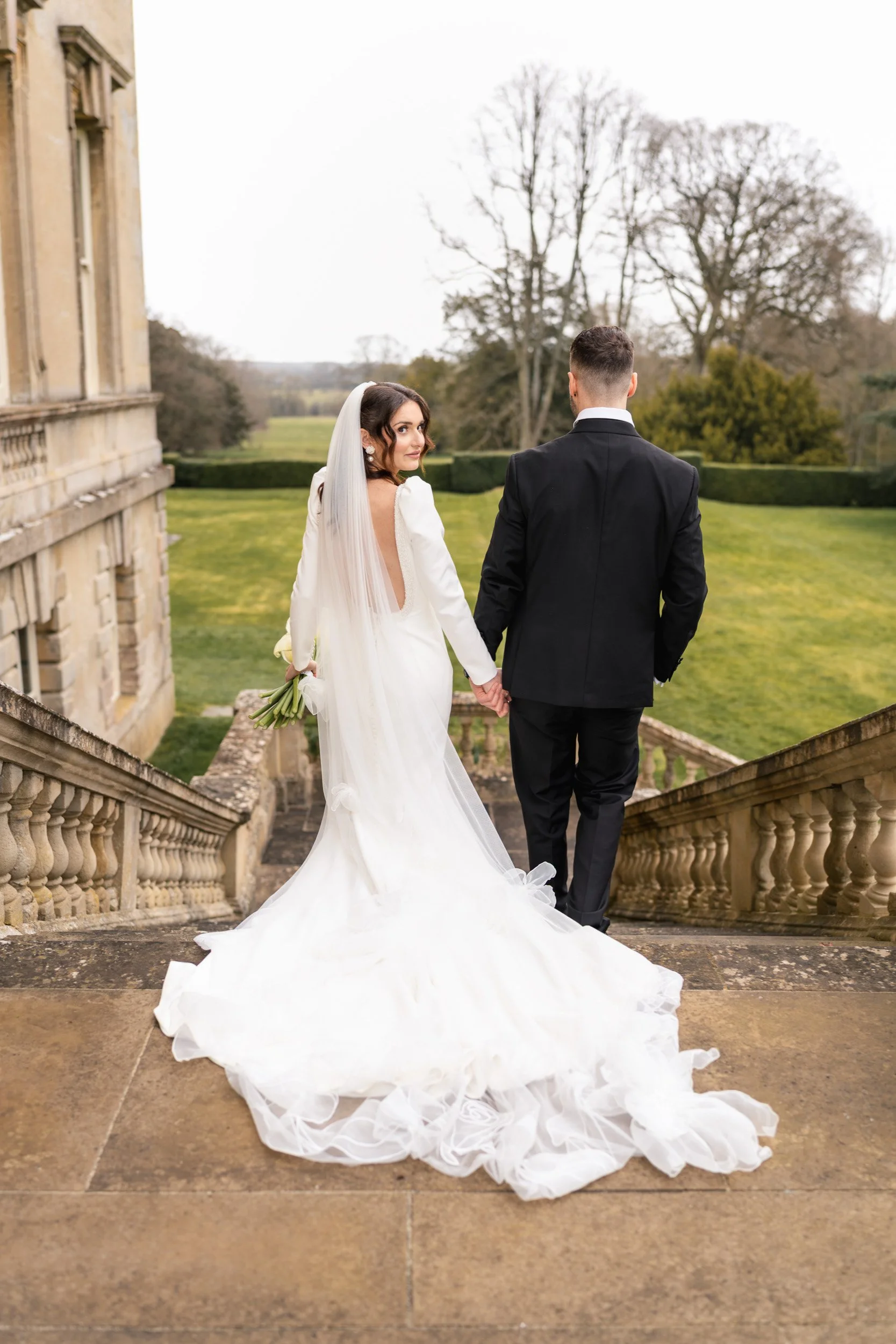 Bride and groom on the garden steps at Kirtlington Park in Oxfordshire with her looking over her shoulder at her wedding guests