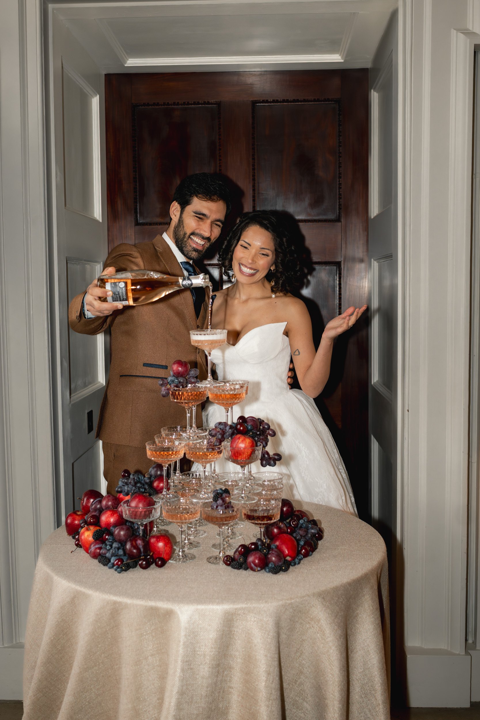 Bride and groom pouring champagne at a fruit and champagne tower during their reception  at Kirtlington Park in Oxfordshire