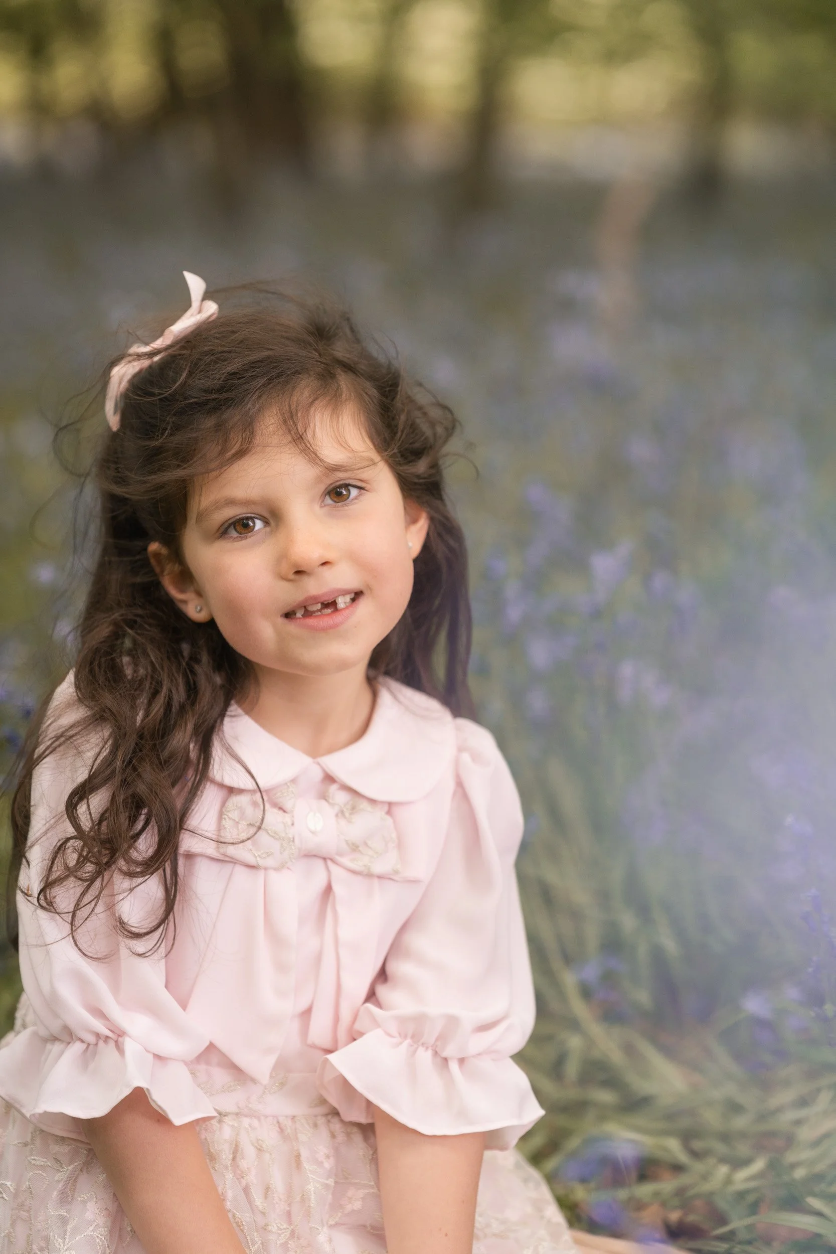 A little girl in a pink dress smiles amongst the flowers  during bluebell season in the Chiltern Hills  near Amersham, Buckinghamshire during a family photography session 