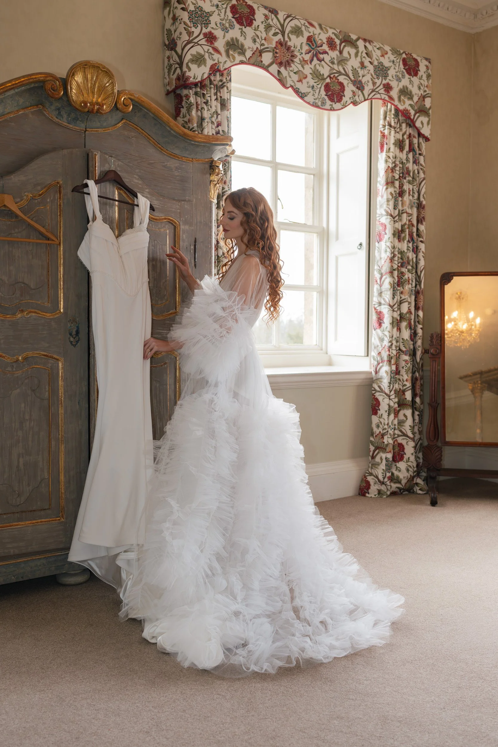 Bride in bridal lingerie and wedding gown touching her wedding dress in the bridal suite at Kirtlington Park in Oxfordshire