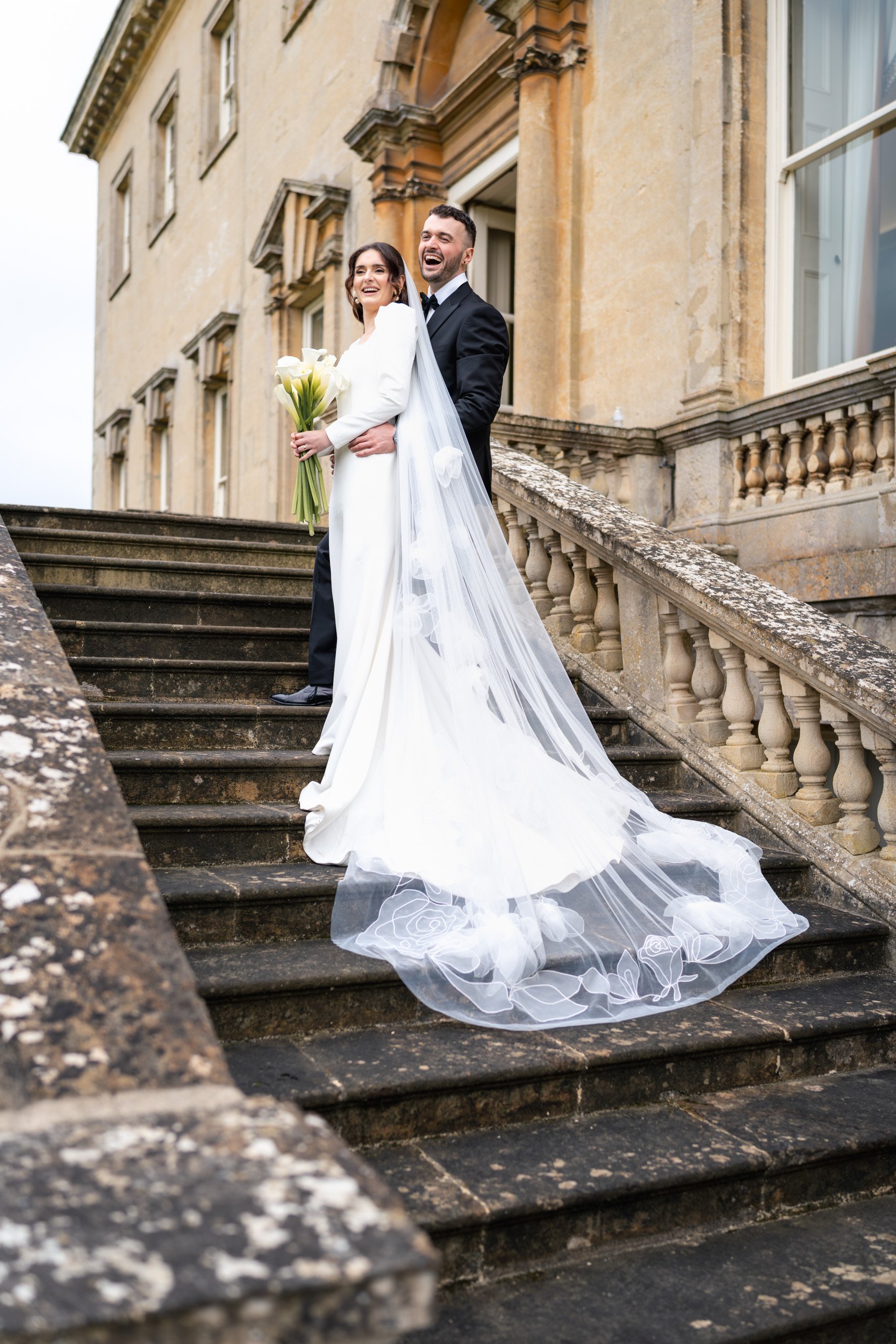 Bride and groom pausing to laugh halfway down the stairs to greet their guests at Kirtlington Park in Oxfordshire