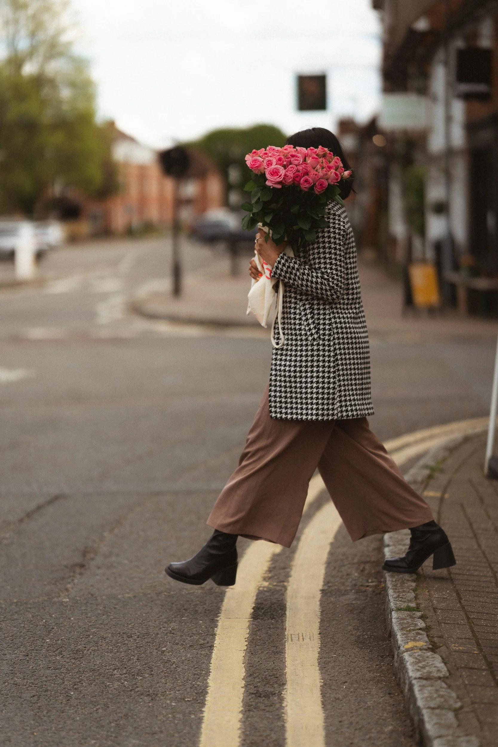 Social media influencer walking across a street holding pink flowers in Old Amersham, Buckinghamshire during a branding shoot
