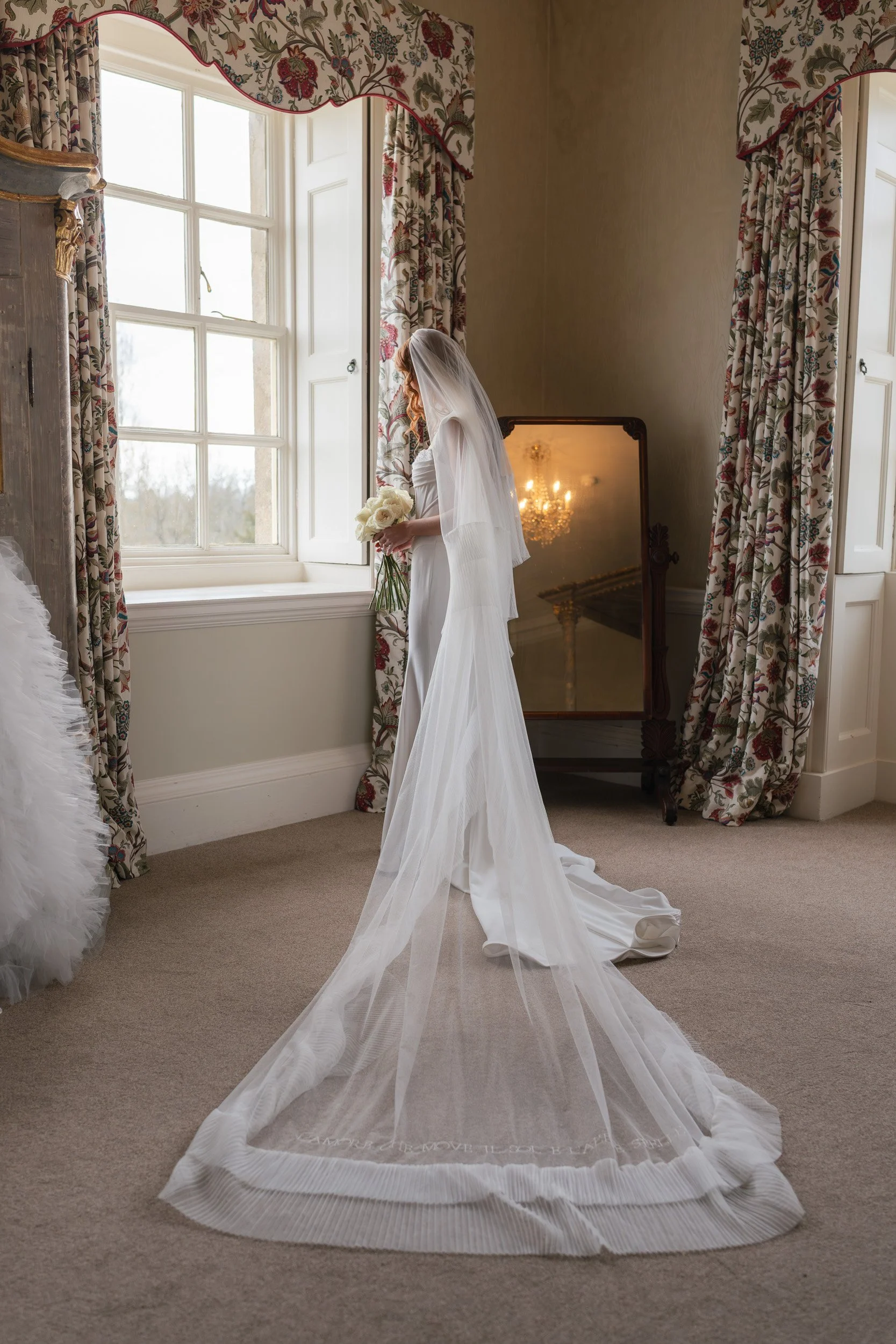 Bride holding her bouquet with her veil at the window in the bridal suite at Kirtlington Park in Oxfordshire