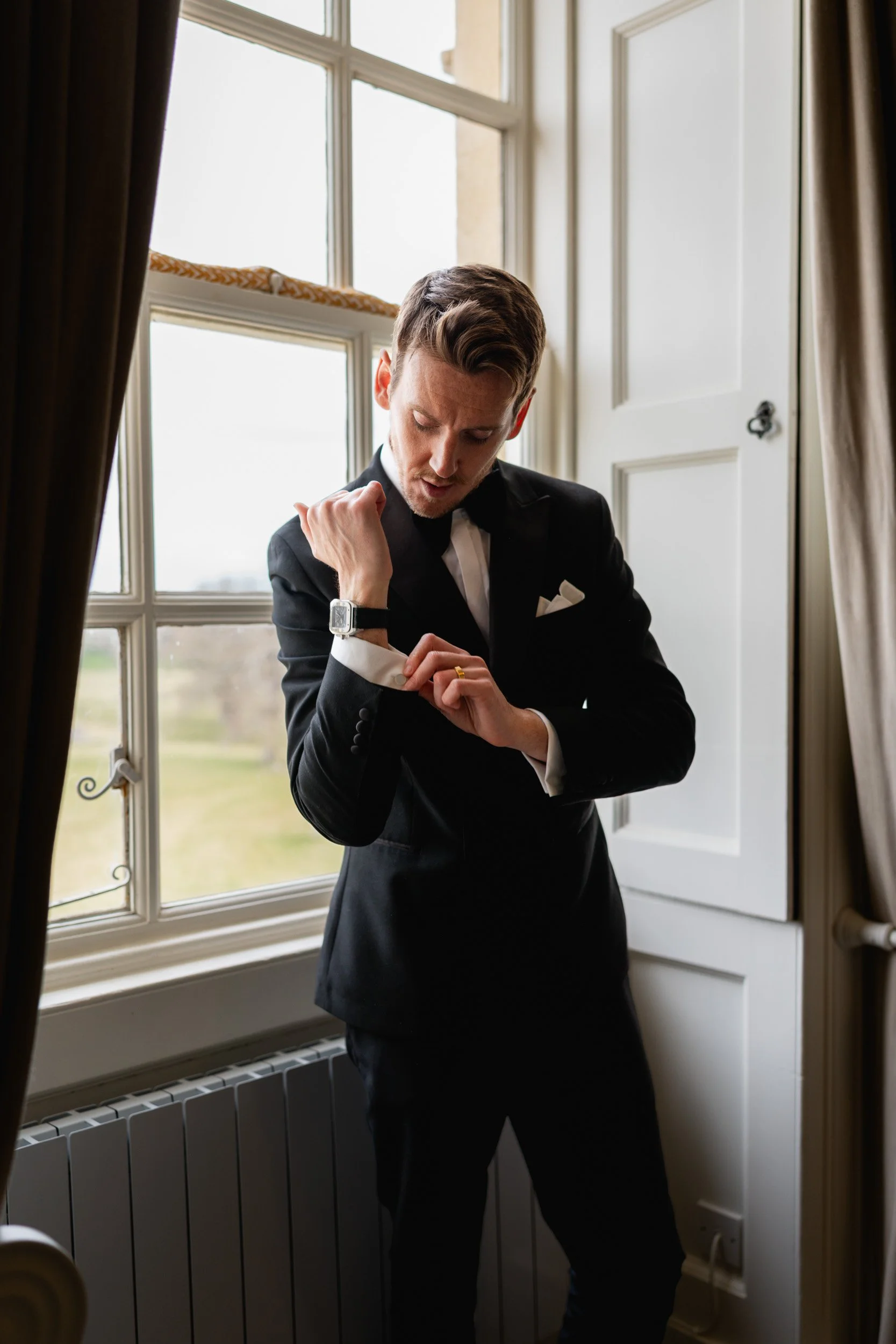 Groom adjusting his cufflinks in the bedroom window as wedding guests arrive at Kirtlington Park in Oxfordshire