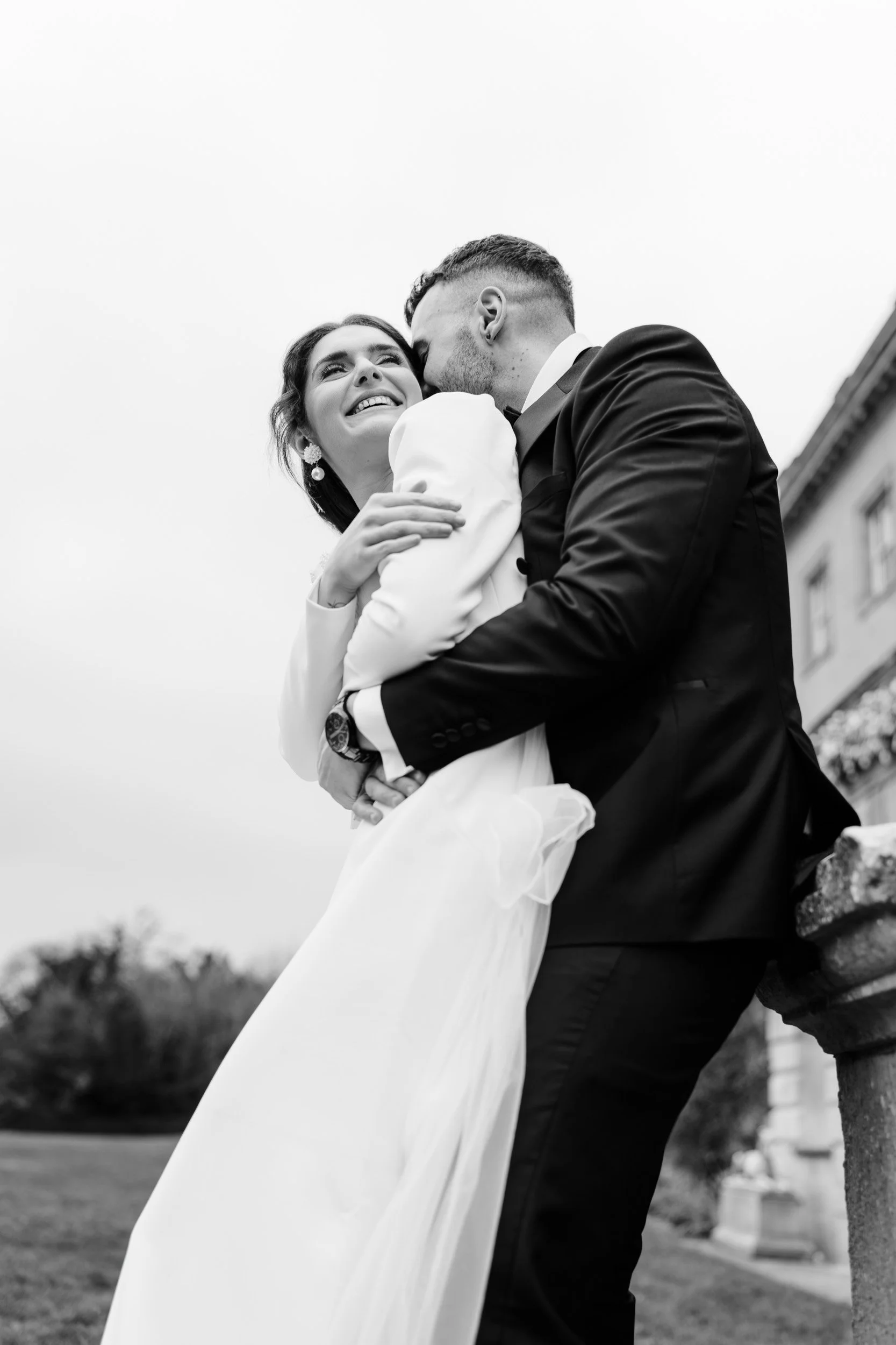 Close up of a groom pulling bride close and whispering in the ear of his bride beside the balustrade in the gardens at Kirtlington Park in Oxfordshire