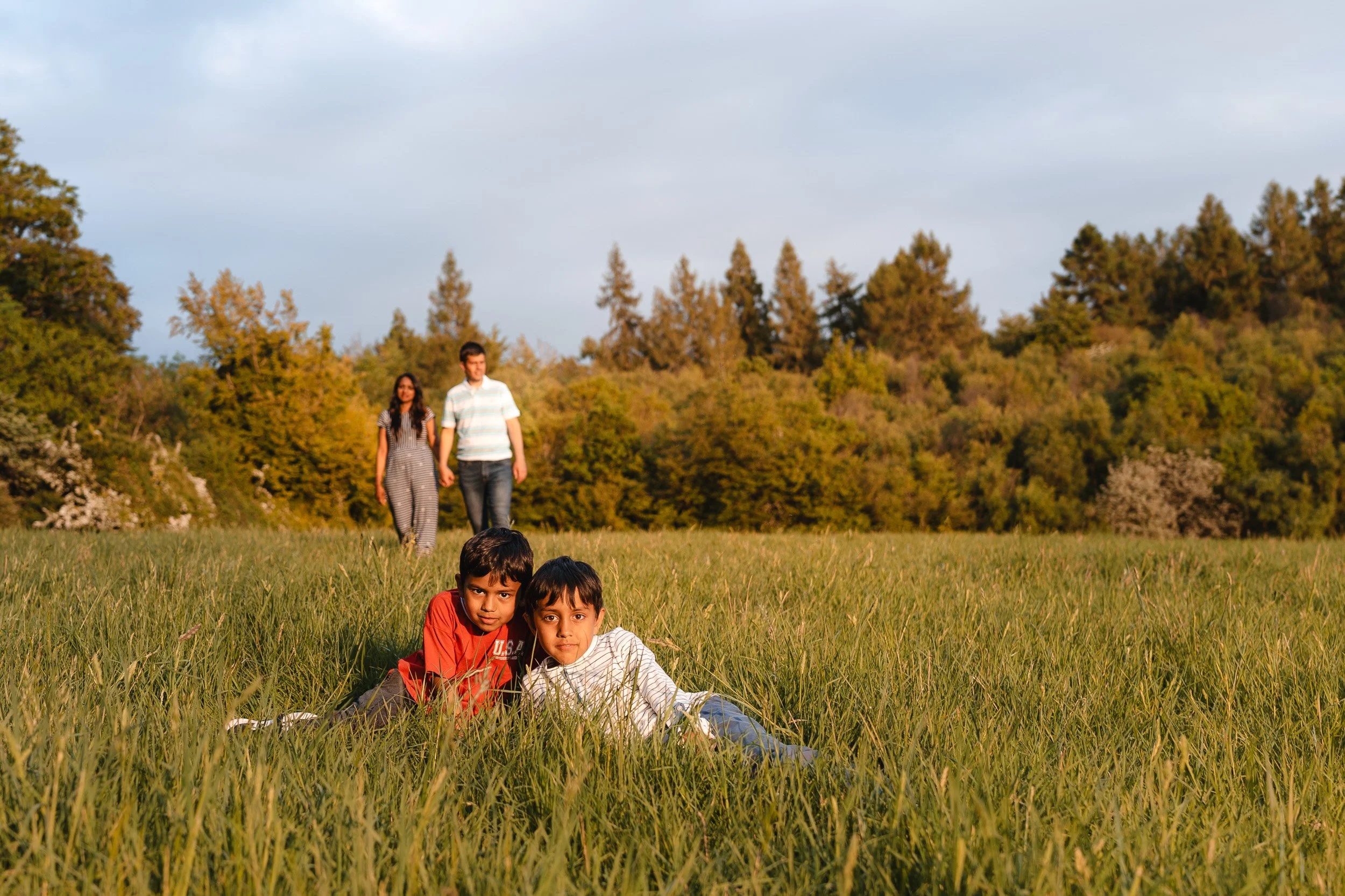 Two young brothers lying in the long grass while their parents walk behind them during a relaxed outdoor family photography session in Amersham, Buckinghamshire 